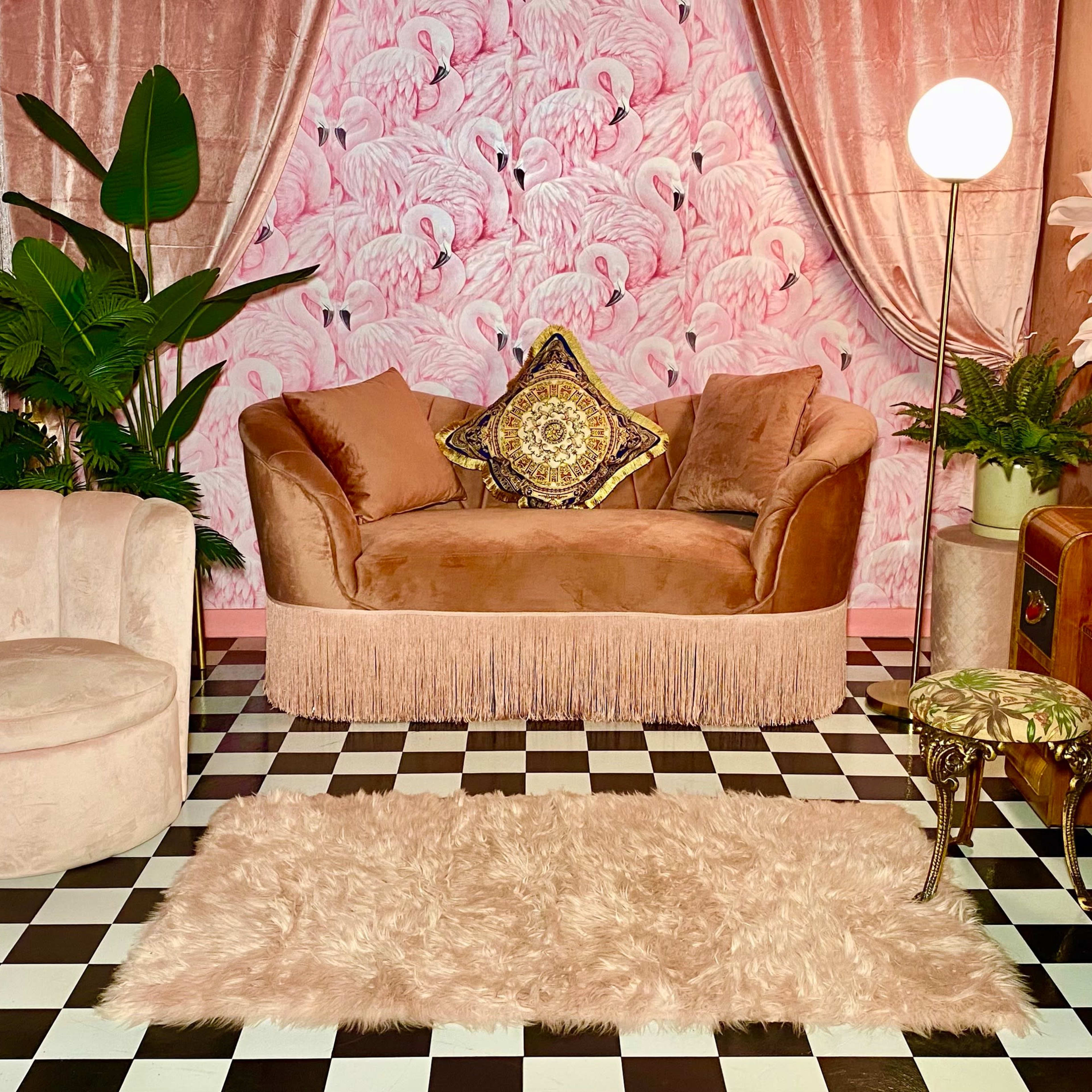A softly lit lounge area features a pink velvet sofa, a round chair, a black and white checkered floor, and a decorative wall with a flamingo pattern.