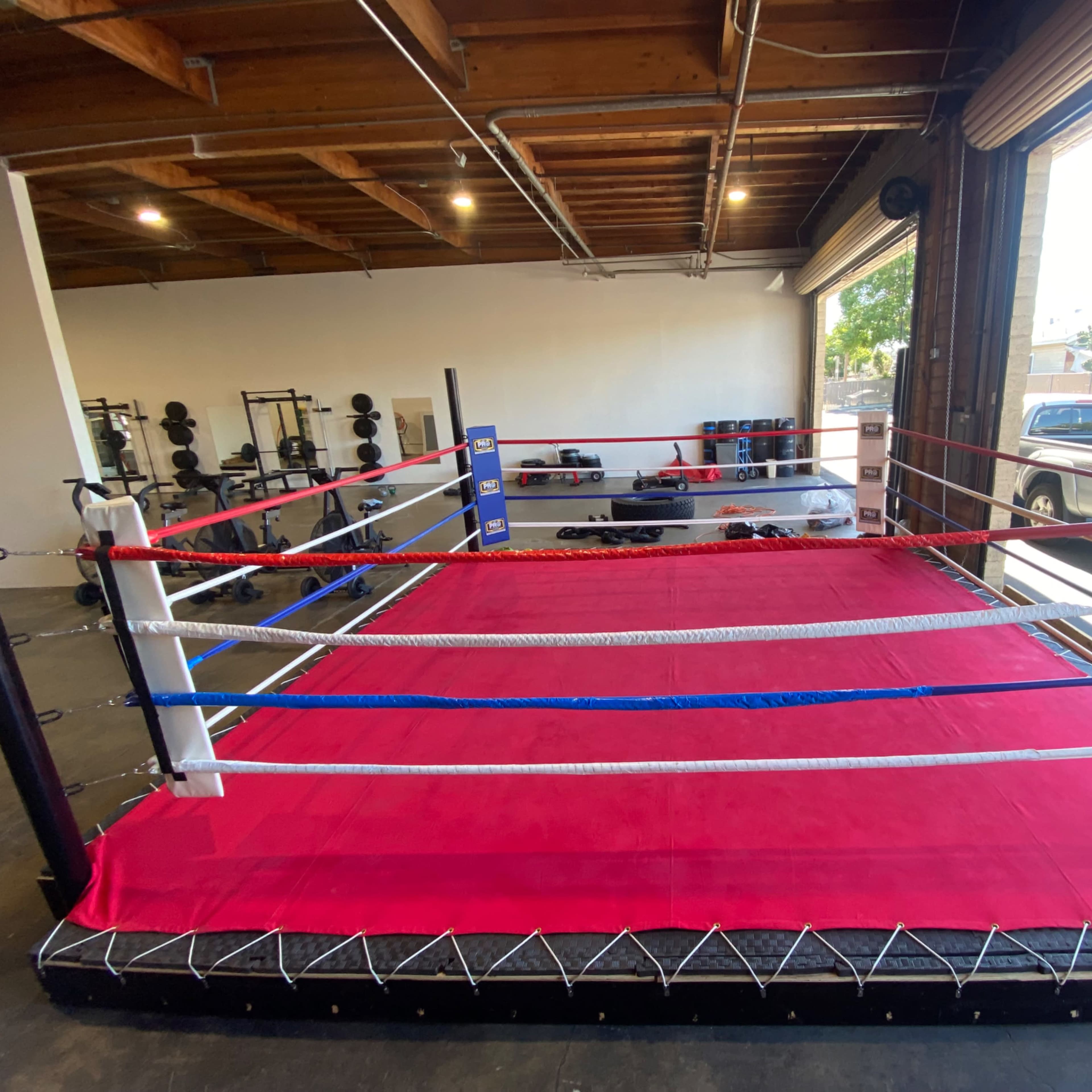 A boxing ring with red and blue corner pads set up in a gym with weightlifting equipment in the background.