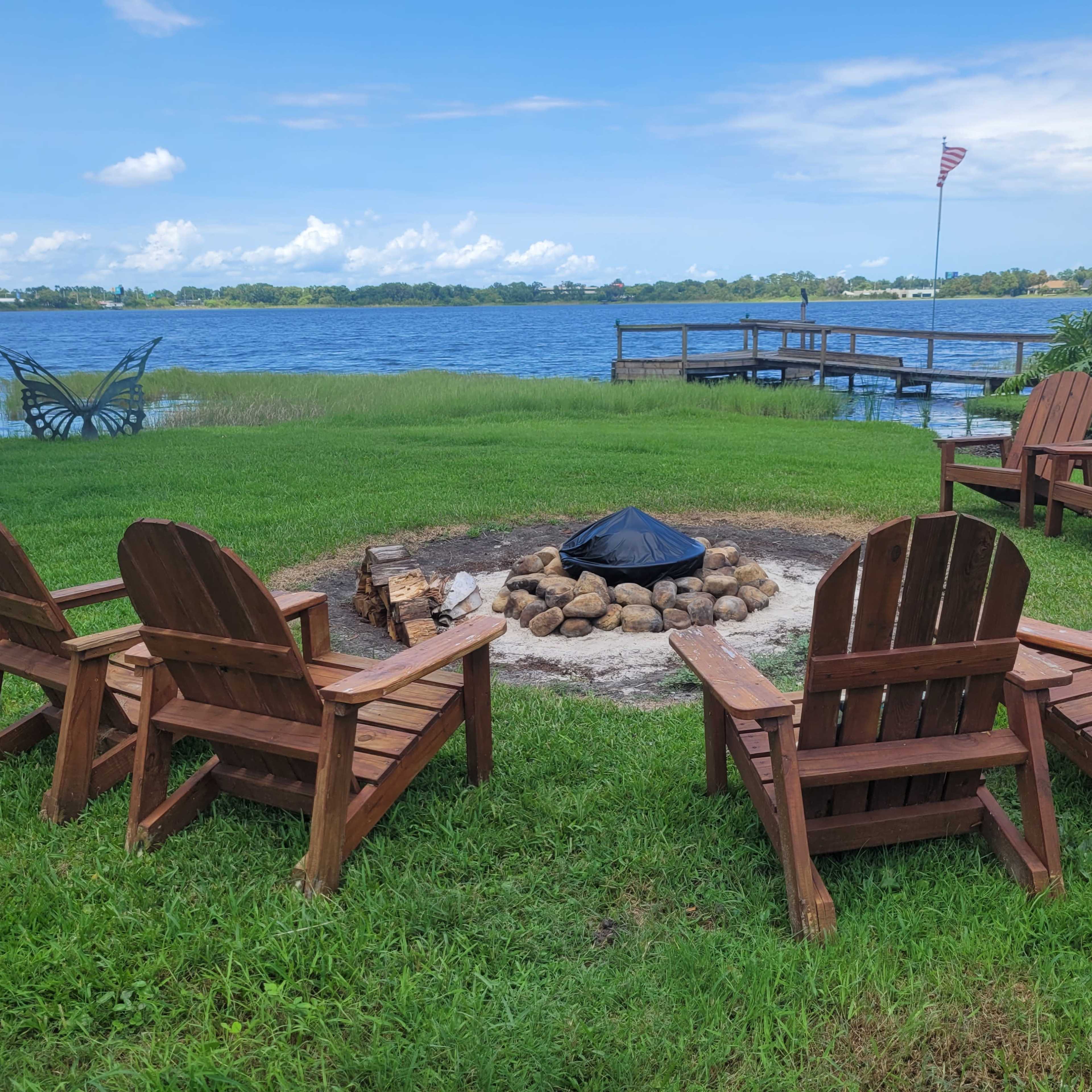 A circular fire pit surrounded by wooden chairs is located by a lake with a dock visible in the background.