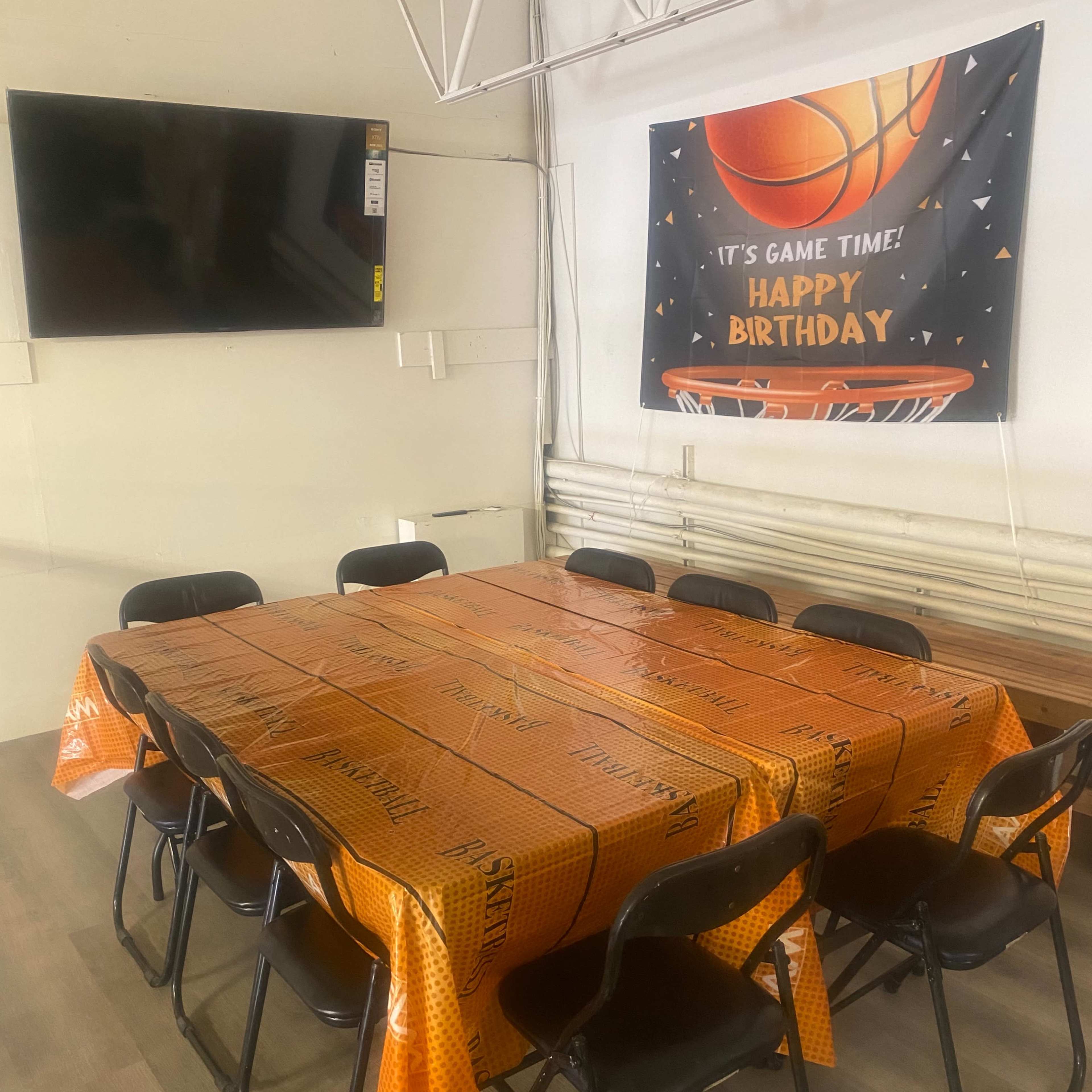 A rectangular table covered with an orange basketball-themed tablecloth is surrounded by black chairs in a decorated room featuring a "Happy Birthday" banner and a television on the wall.