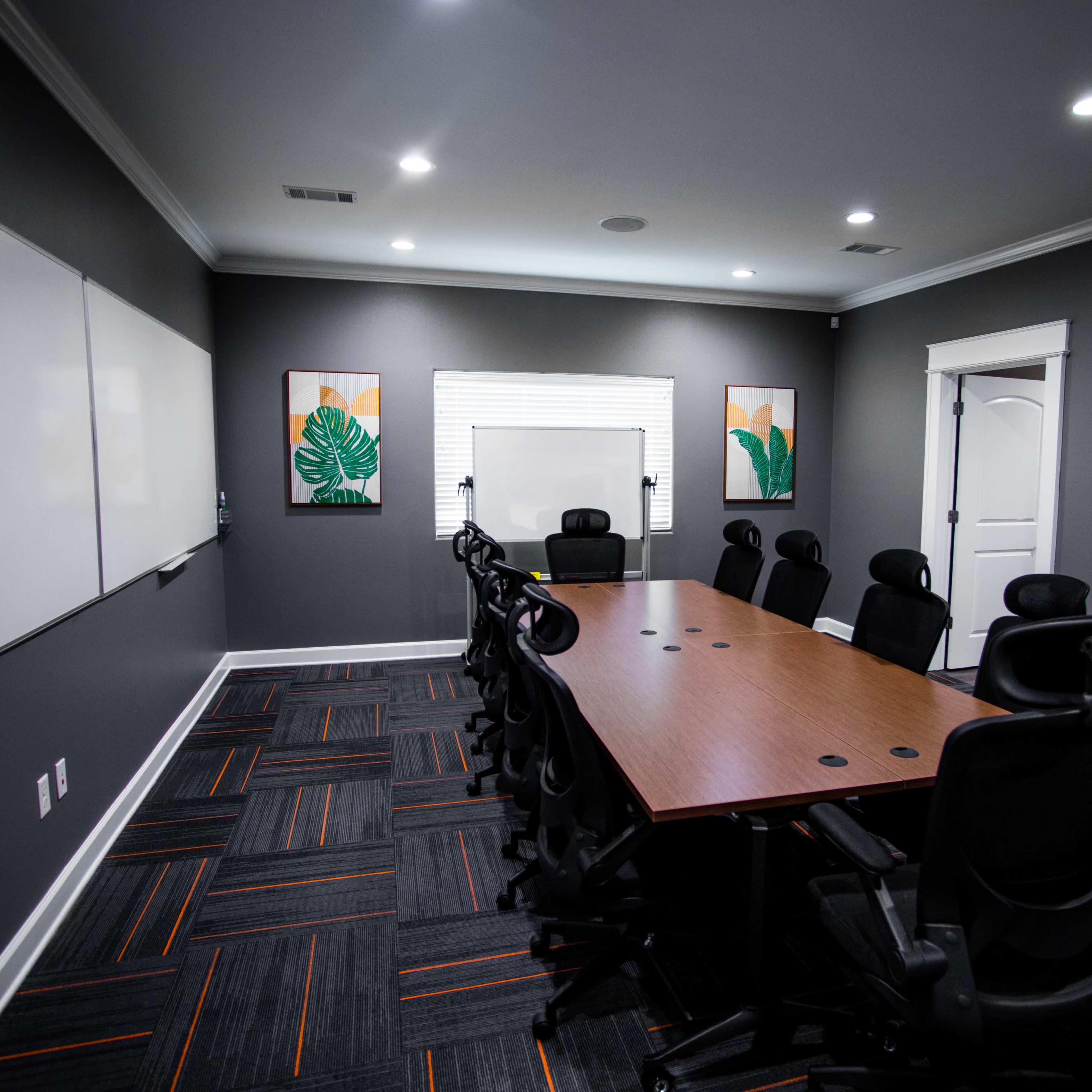 A modern conference room featuring a long wooden table surrounded by black office chairs, with whiteboards on the walls and framed leaf prints hanging decoratively.