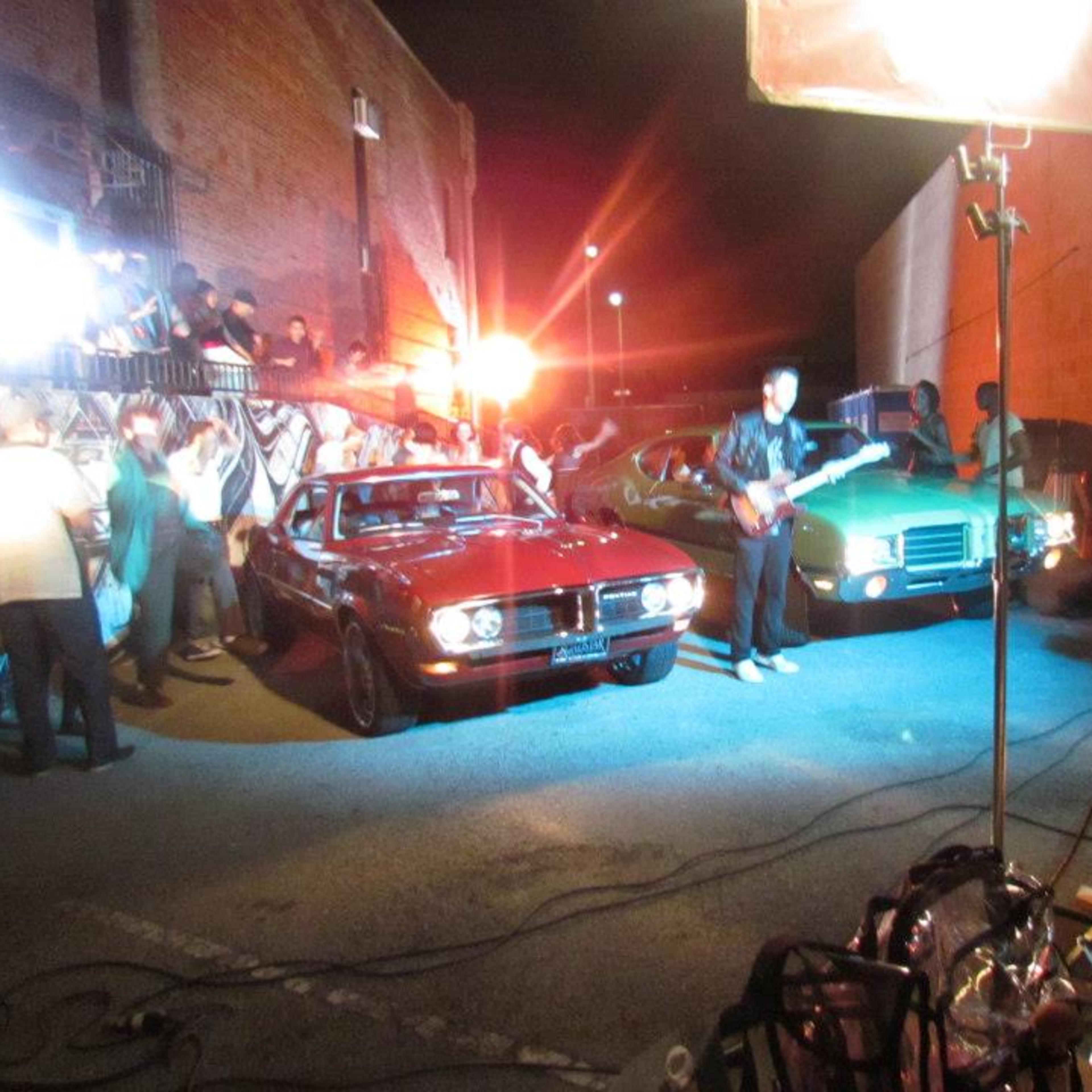 A film crew sets up a night shoot featuring two classic cars, surrounded by people and lights in an alley.