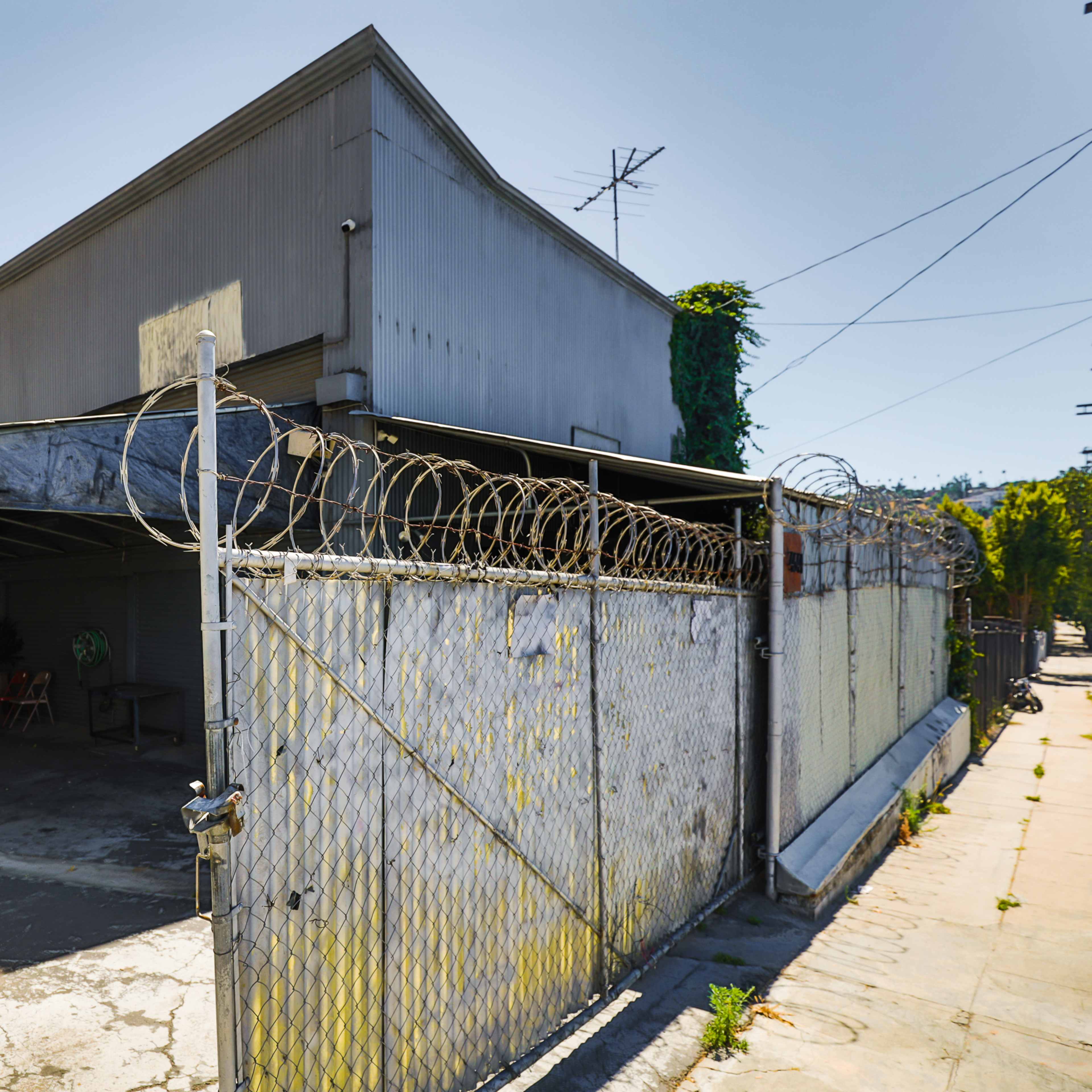 A metal fence topped with barbed wire surrounds a gray building with a flat roof and an empty driveway, set against a clear blue sky.