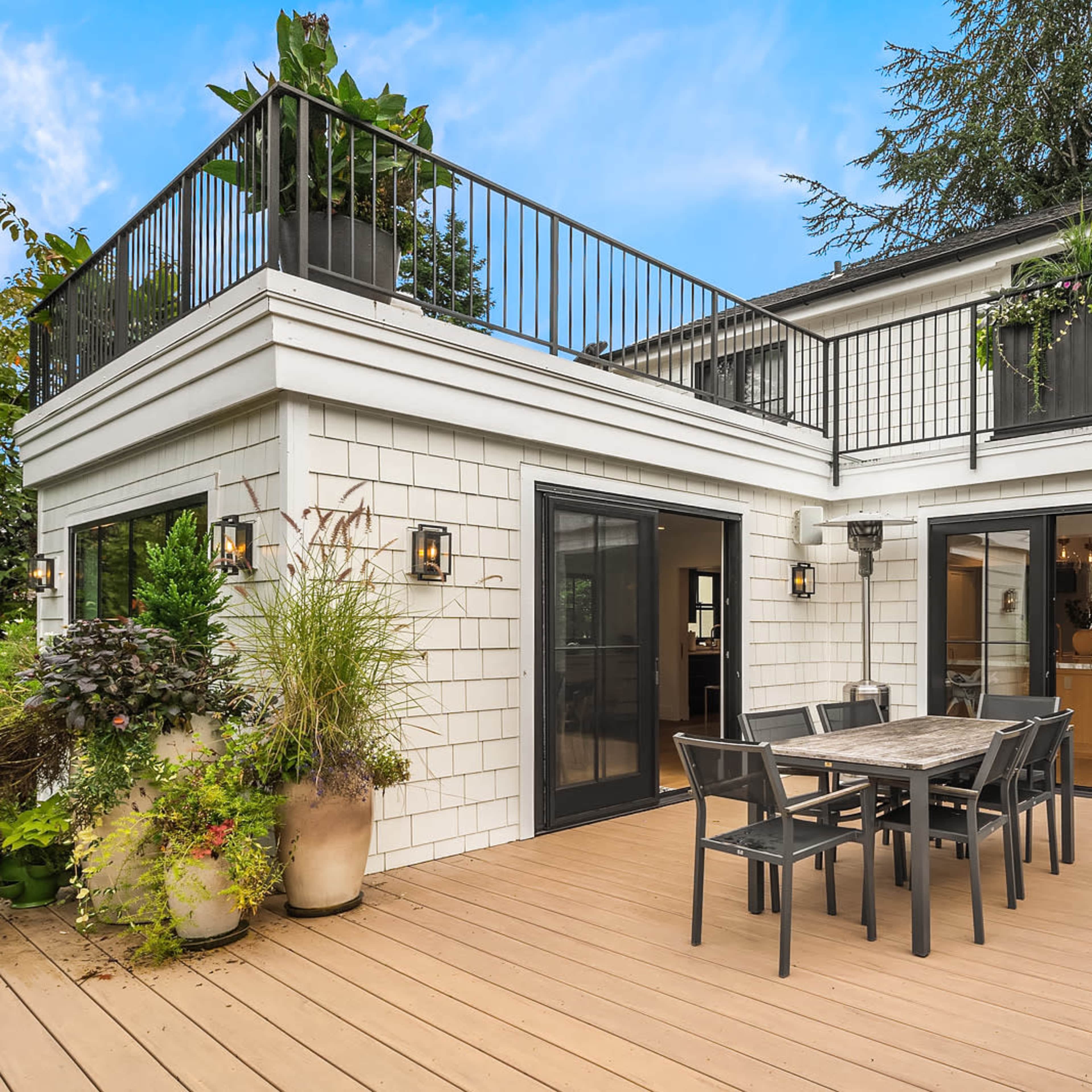 A modern outdoor dining area on a wooden deck, featuring a table with chairs, a grill, and potted plants, adjacent to a white brick building with black-framed glass doors.