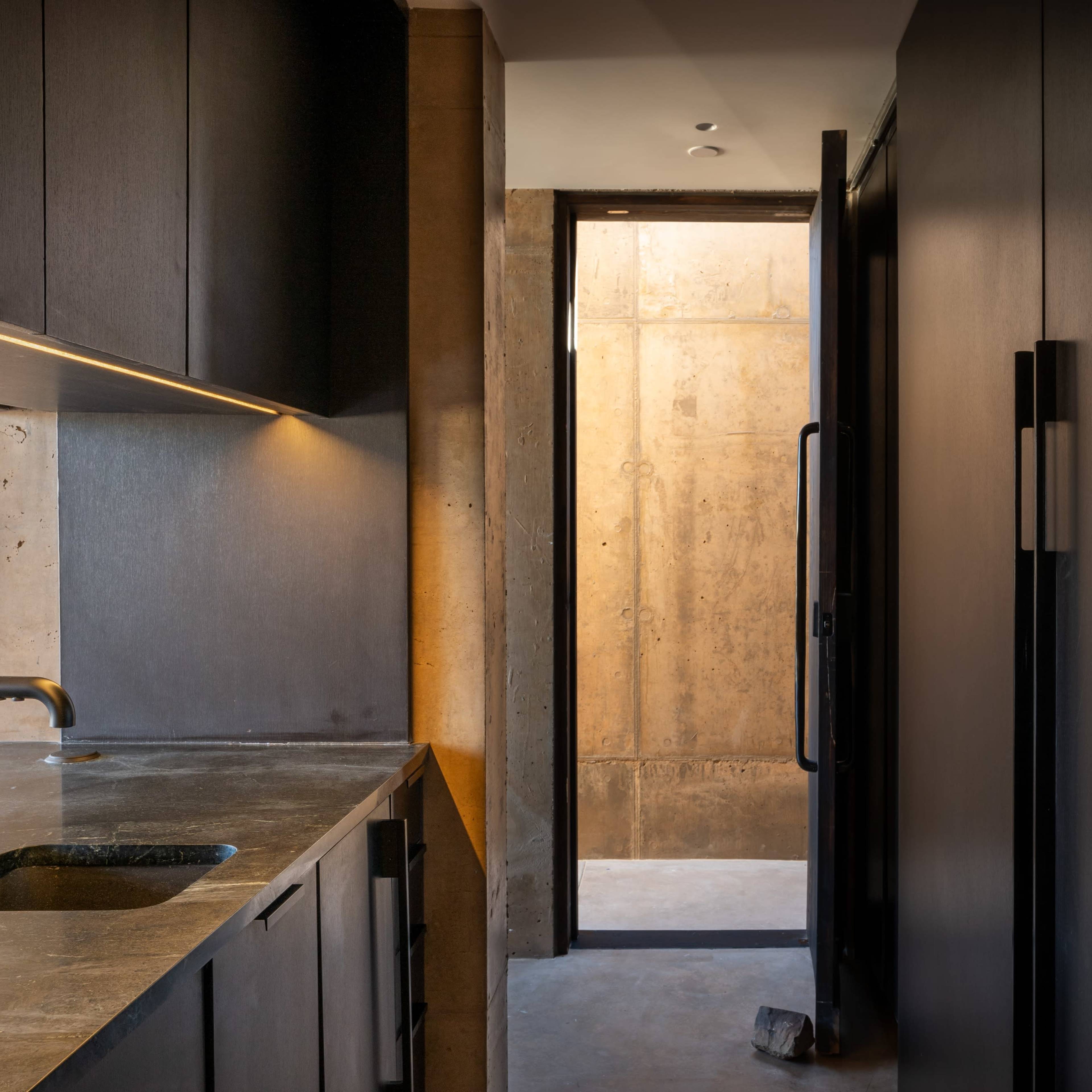 A narrow kitchen space with dark cabinetry and a doorway leading to a light-filled hallway with concrete walls.