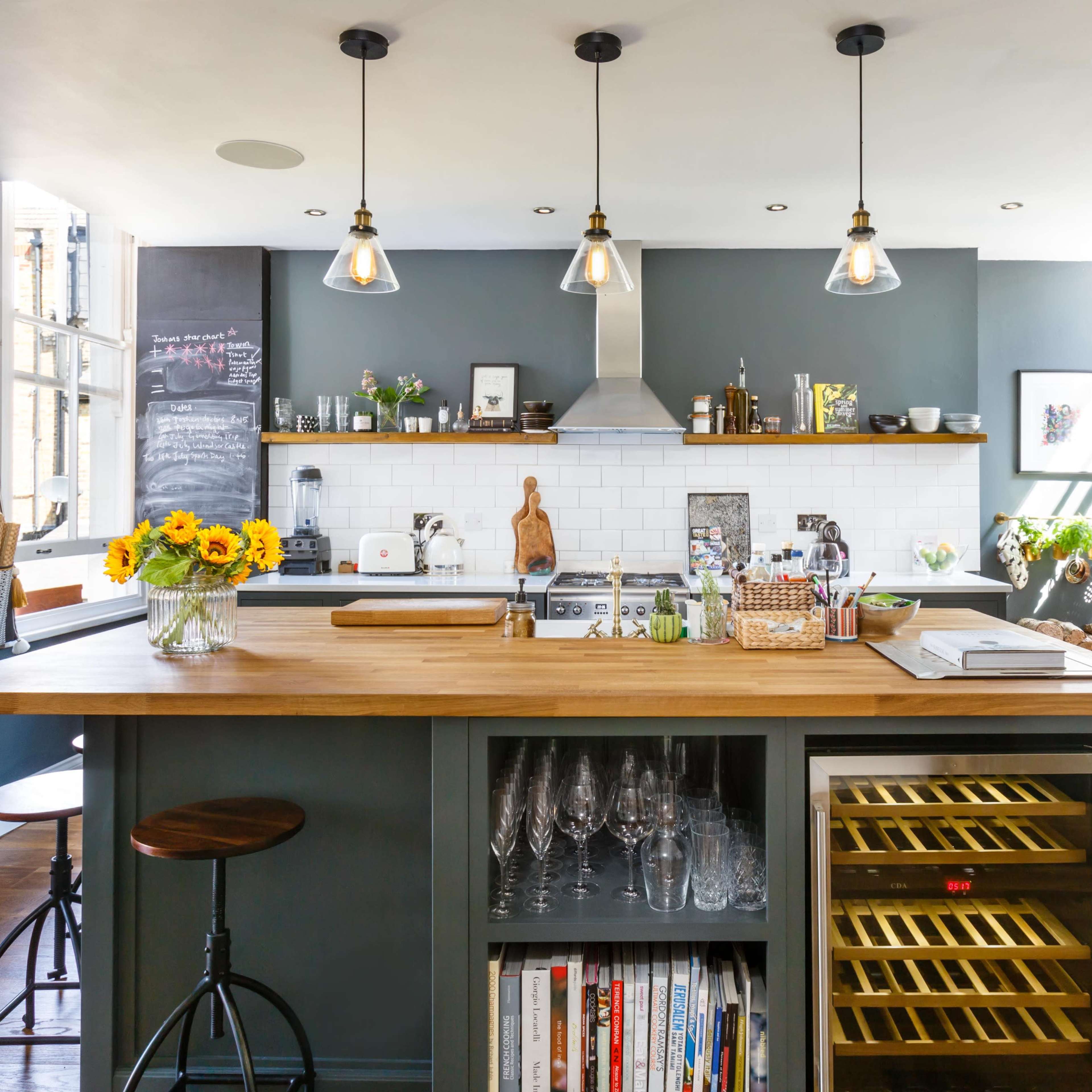 A modern kitchen features a central wooden island with bar stools, a wine fridge, and shelves filled with glassware and cookbooks.