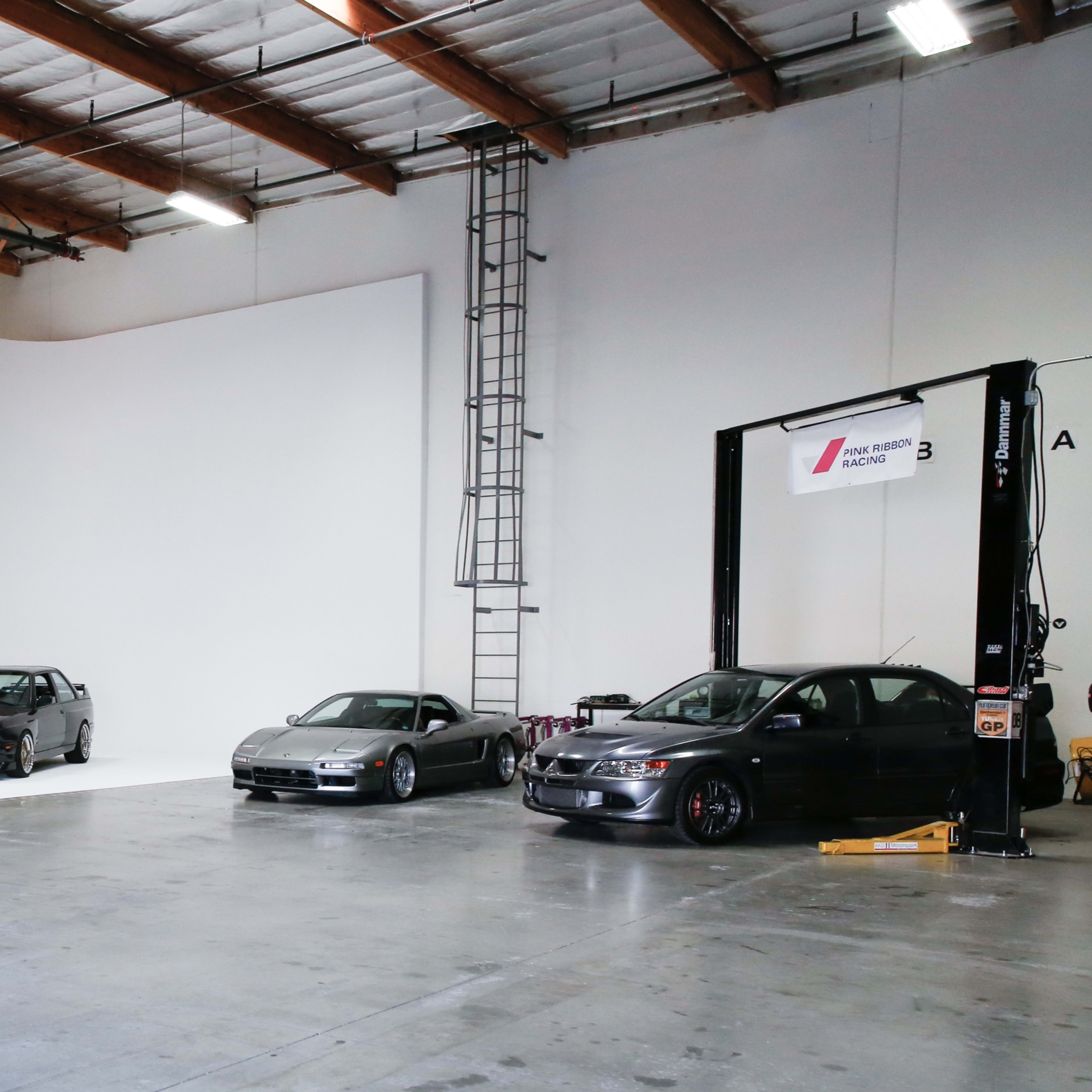 A spacious garage featuring several classic cars, a large white wall, and minimal furnishings, with the words "DEAD SLOW GARAGE" displayed on the wall.