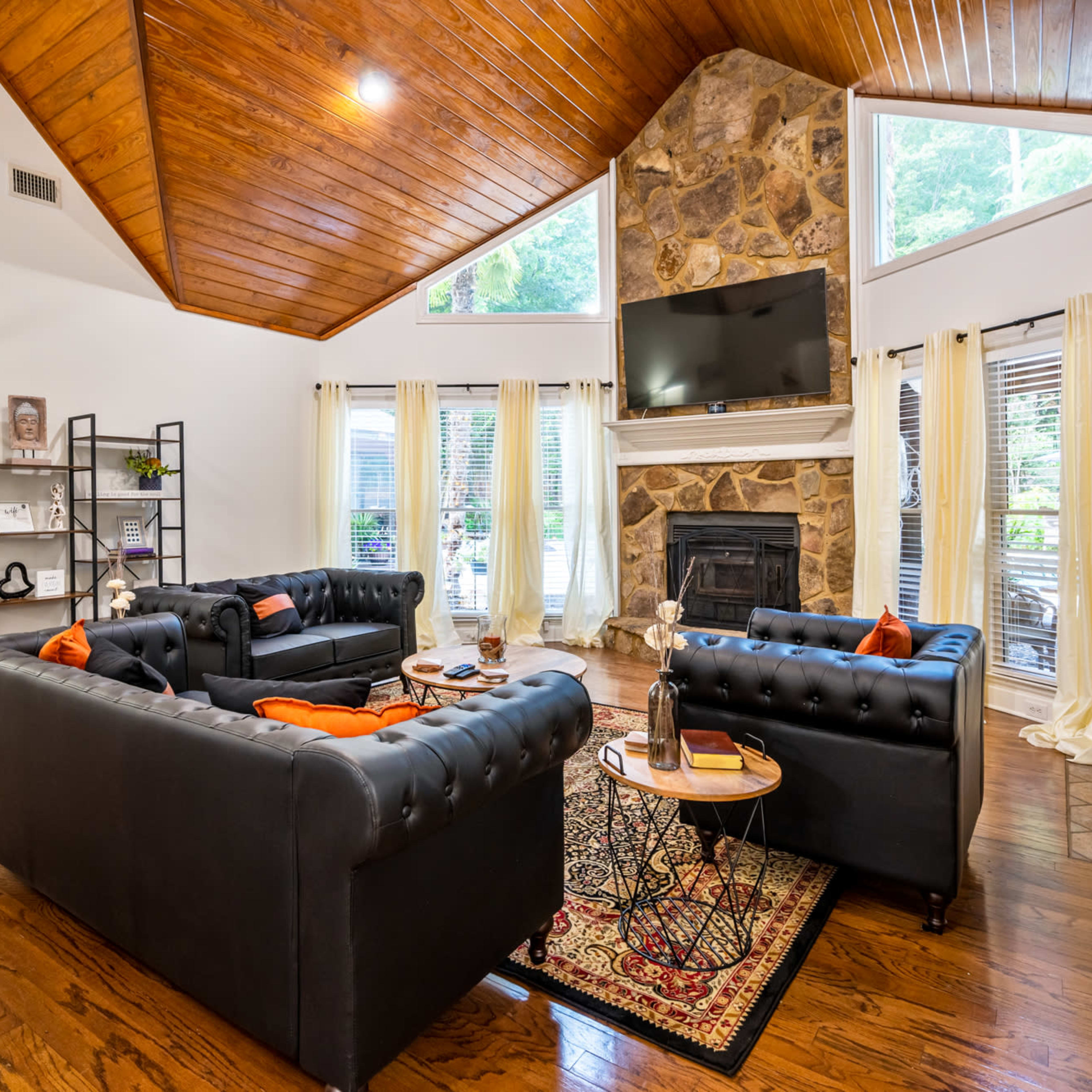 A living room with high wooden ceilings, a stone fireplace, and two dark sofas arranged around a coffee table, with large windows allowing natural light to enter.