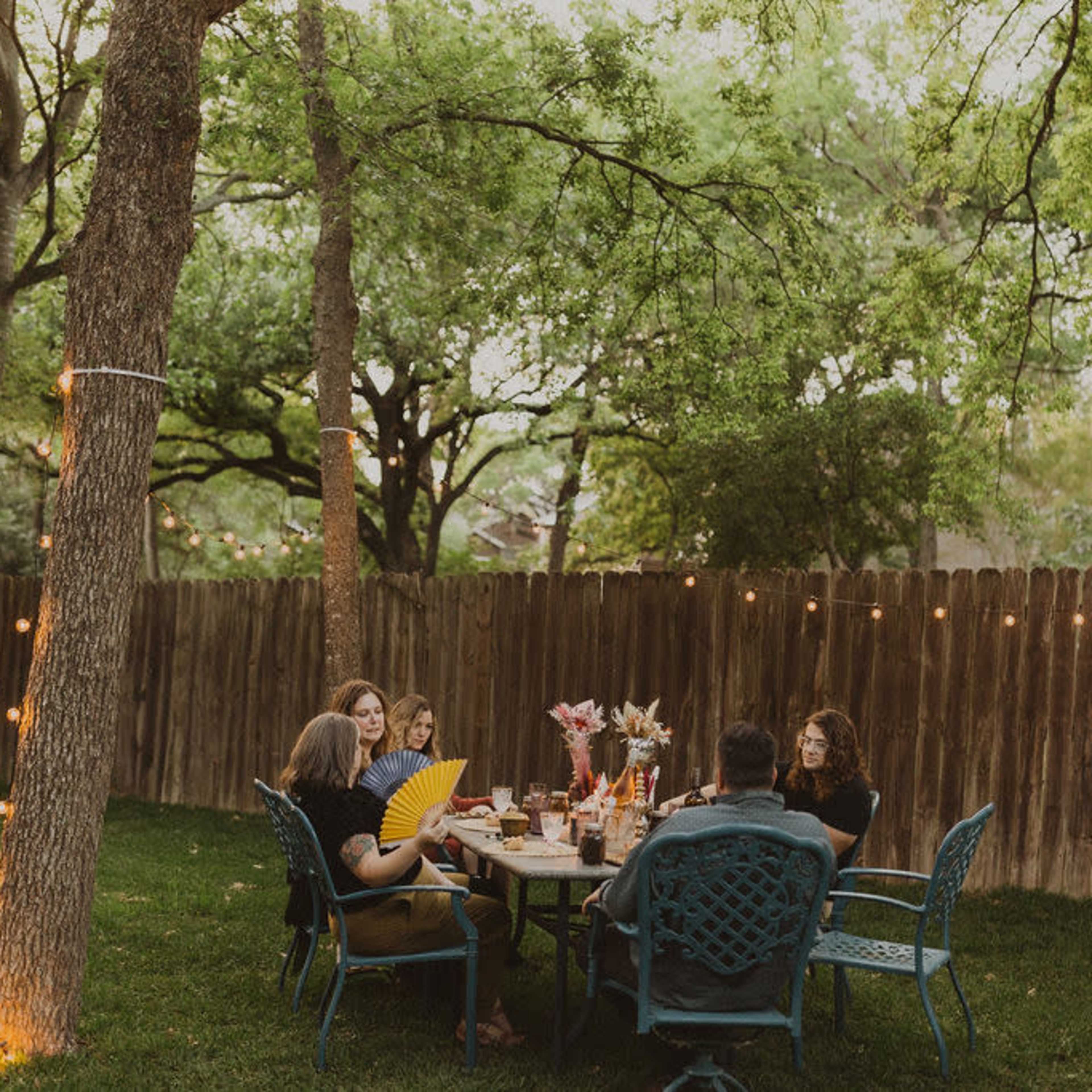 A group of five people is seated around a table in a backyard, with string lights illuminating the scene and trees surrounding the area.