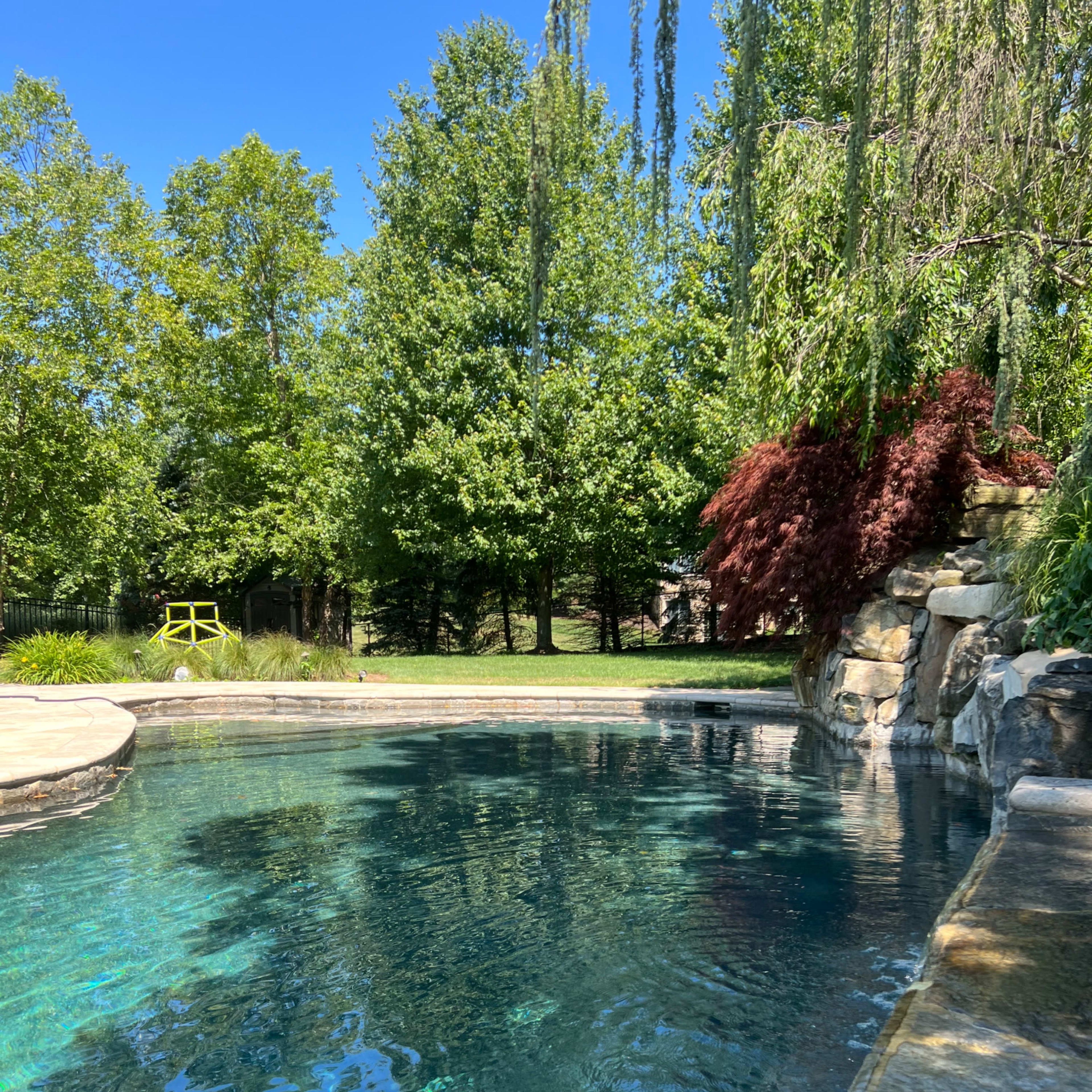 A clear blue swimming pool reflects the surrounding trees and landscape, with a stone edge and a rock formation nearby.