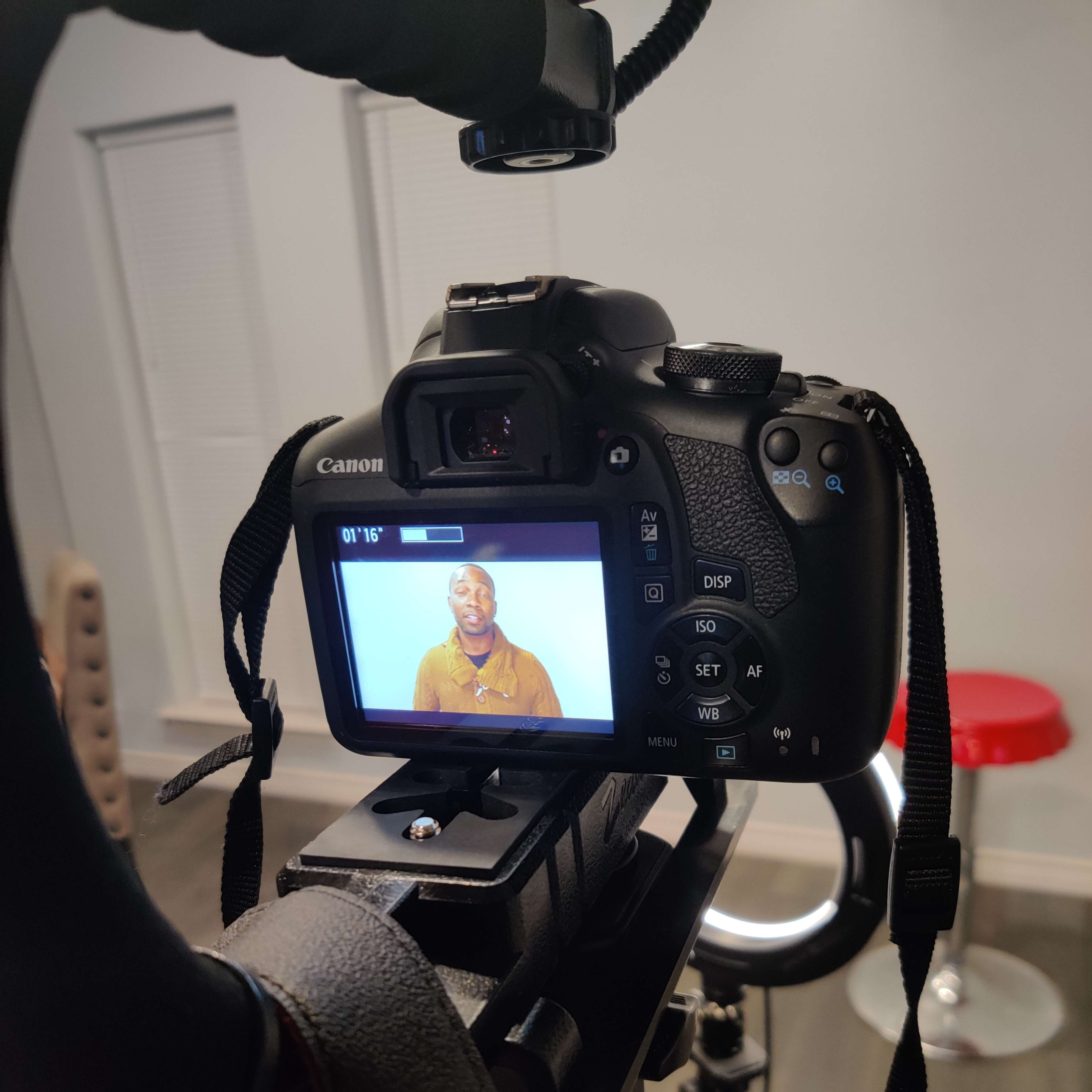 A Canon camera mounted on a tripod, focusing on a person sitting in front of a lighted backdrop, with a red stool visible in the background.
