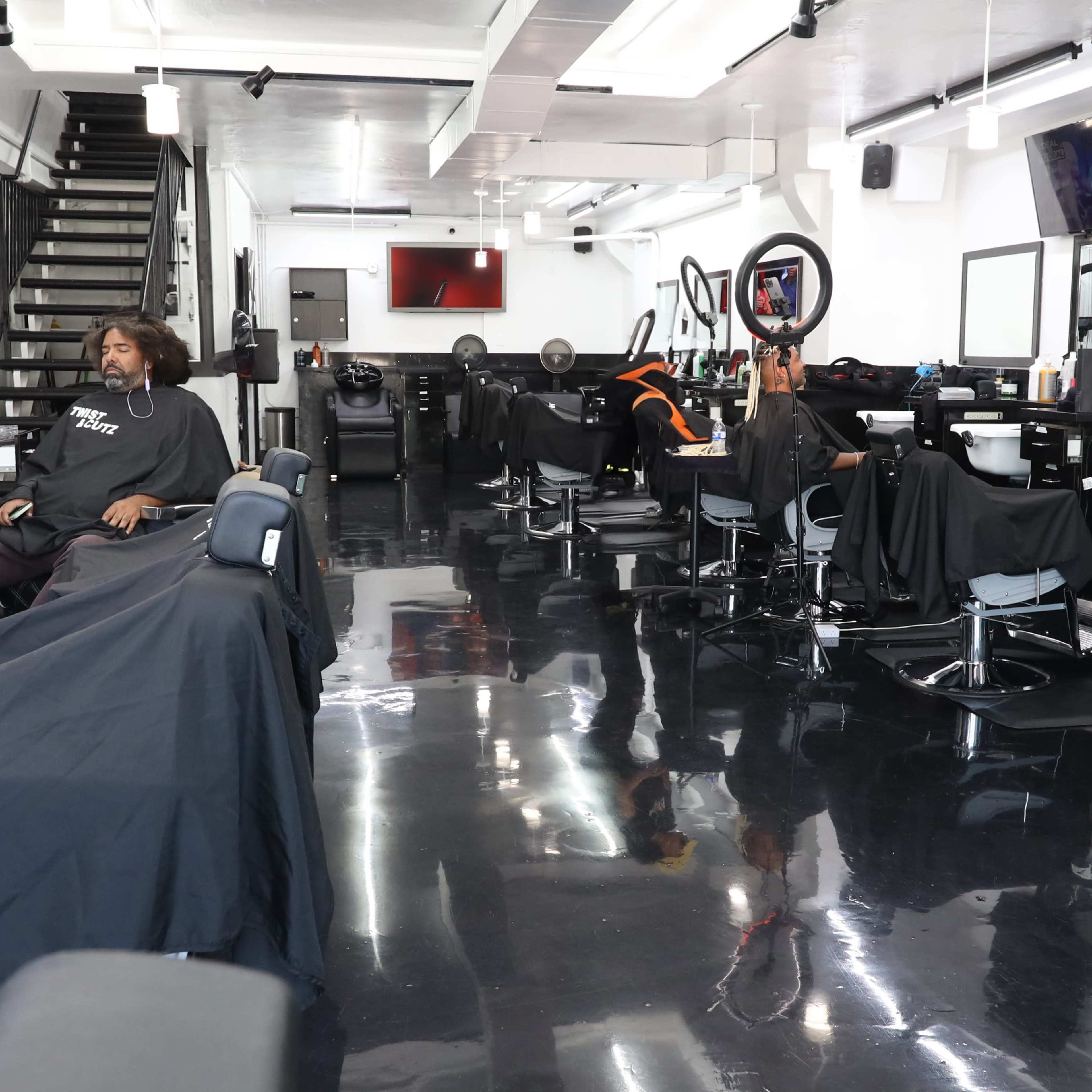 A barber shop with several barber stations, each occupied by clients under black capes, and a staircase in the background.