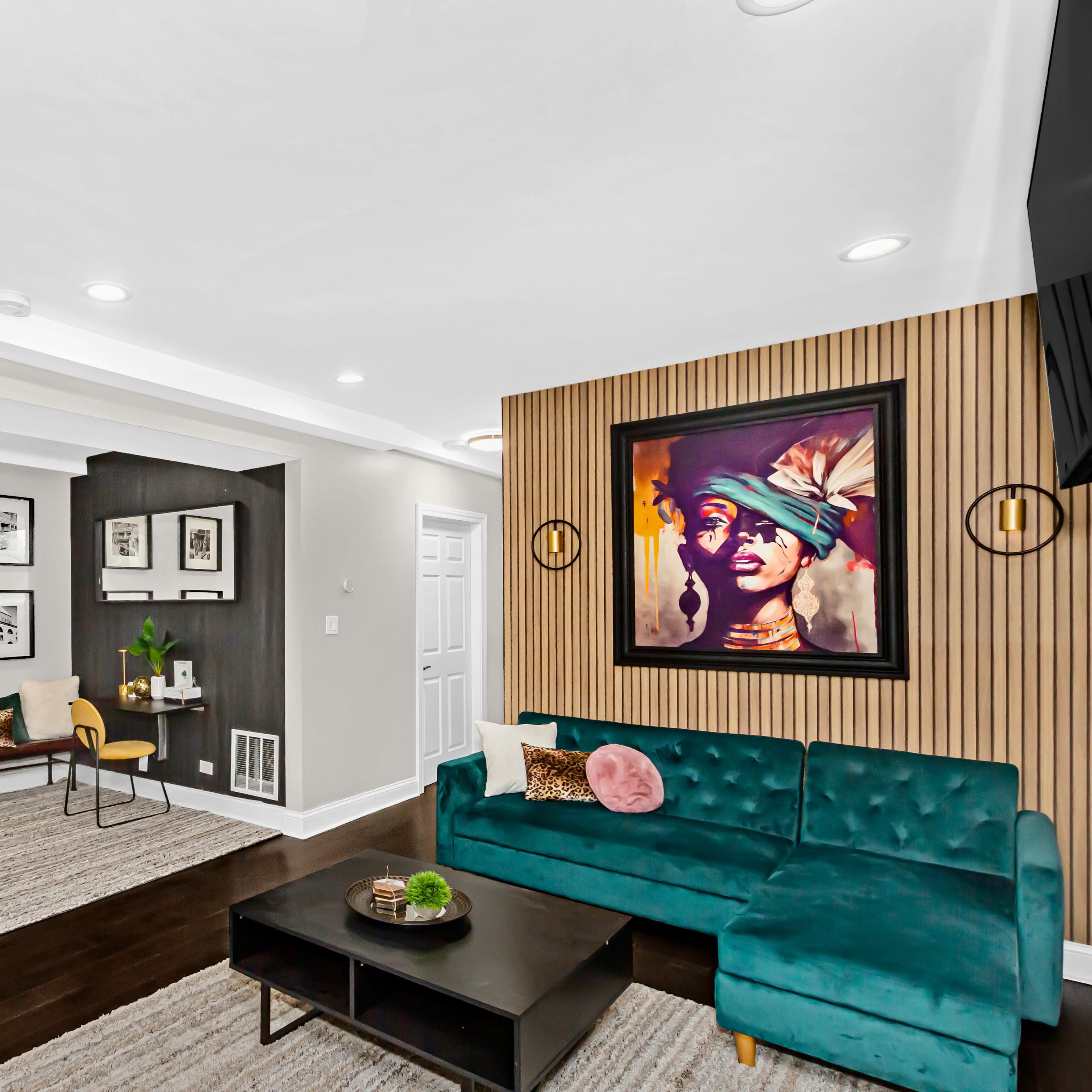 A modern living room with a teal sectional sofa, a wooden bench for seating, framed black-and-white photographs on the walls, and a large colorful artwork above the couch.