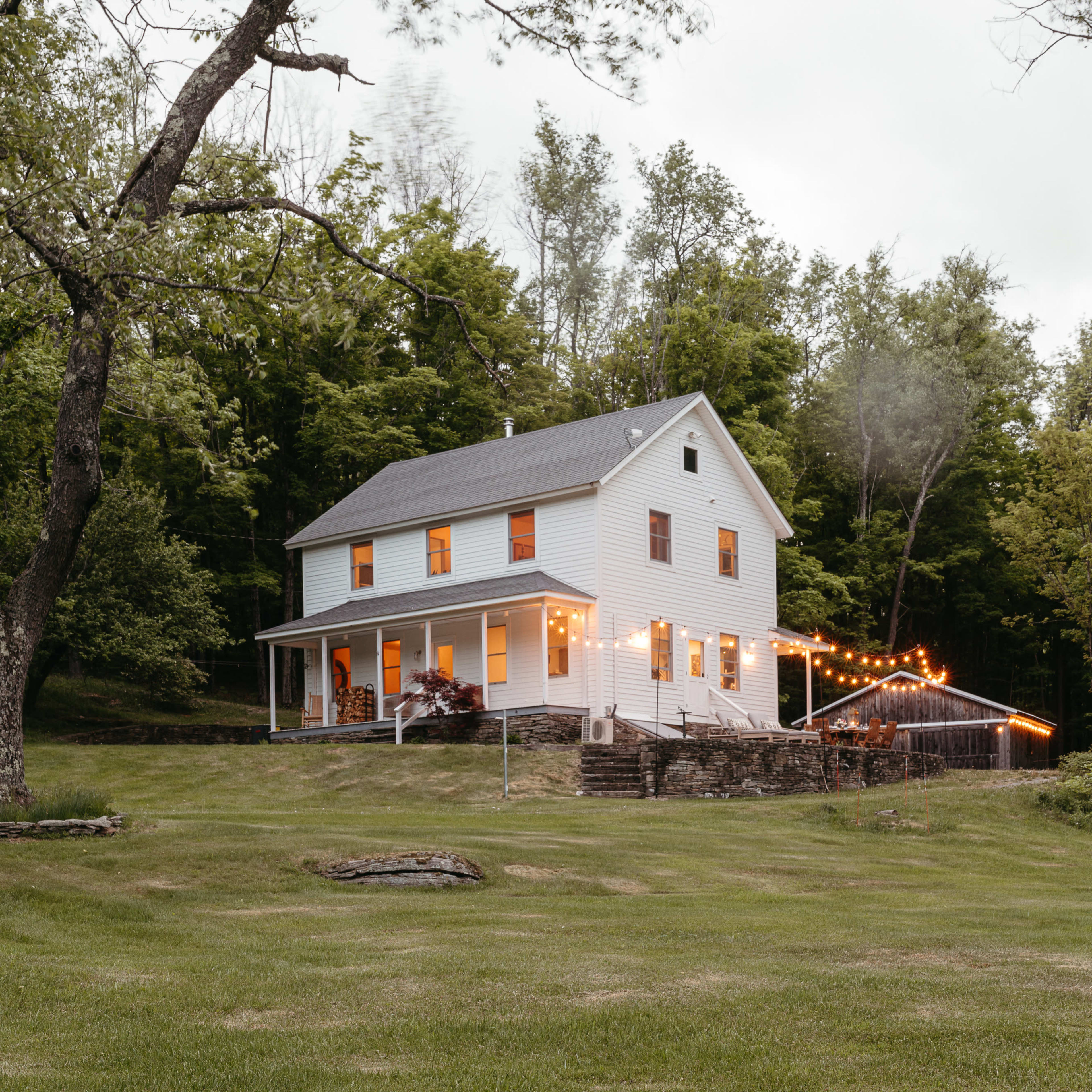 A white two-story house with wooden steps and string lights is set against a backdrop of green trees and an expansive lawn.