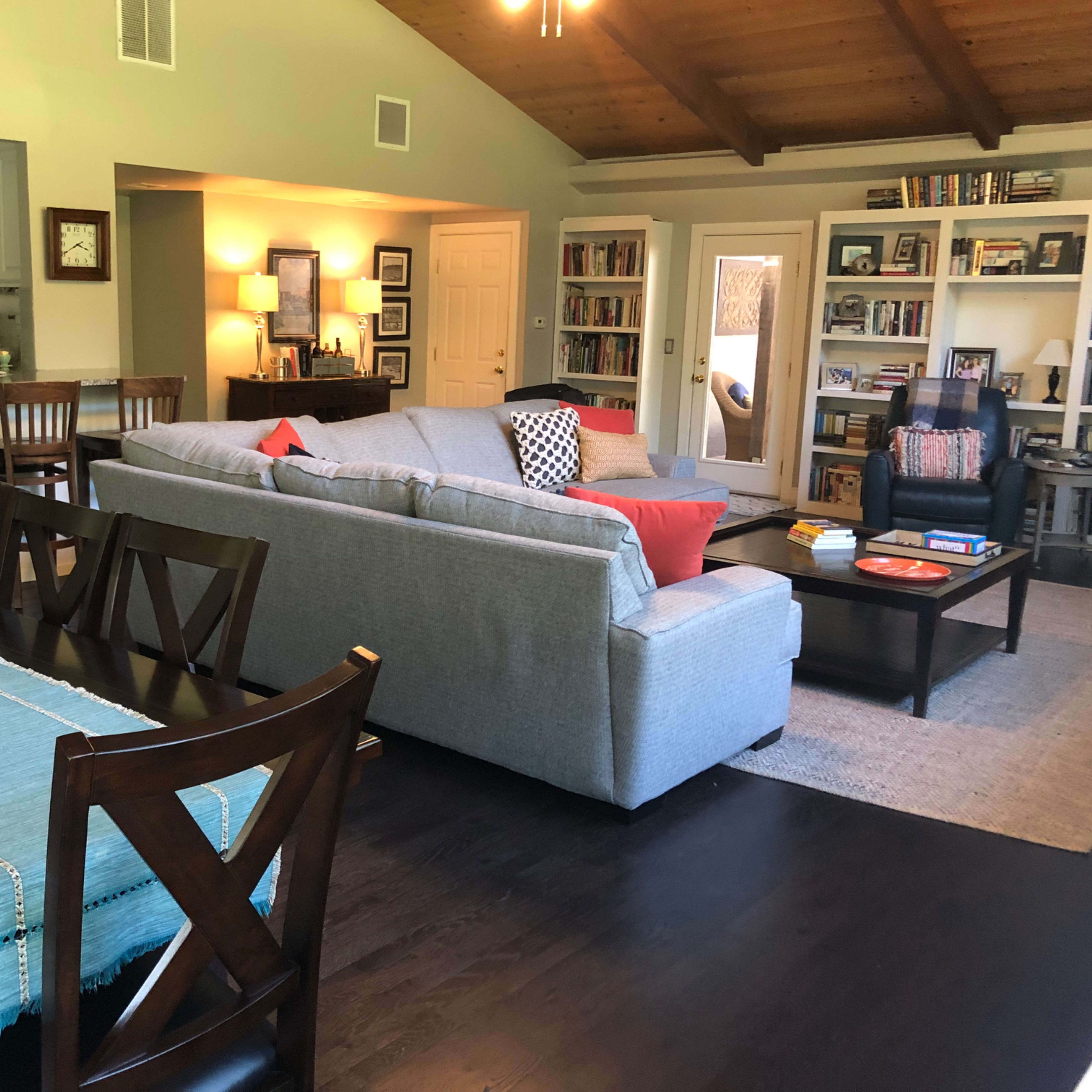 A spacious living room and dining area with a gray sofa, wooden dining table, and shelves filled with books.