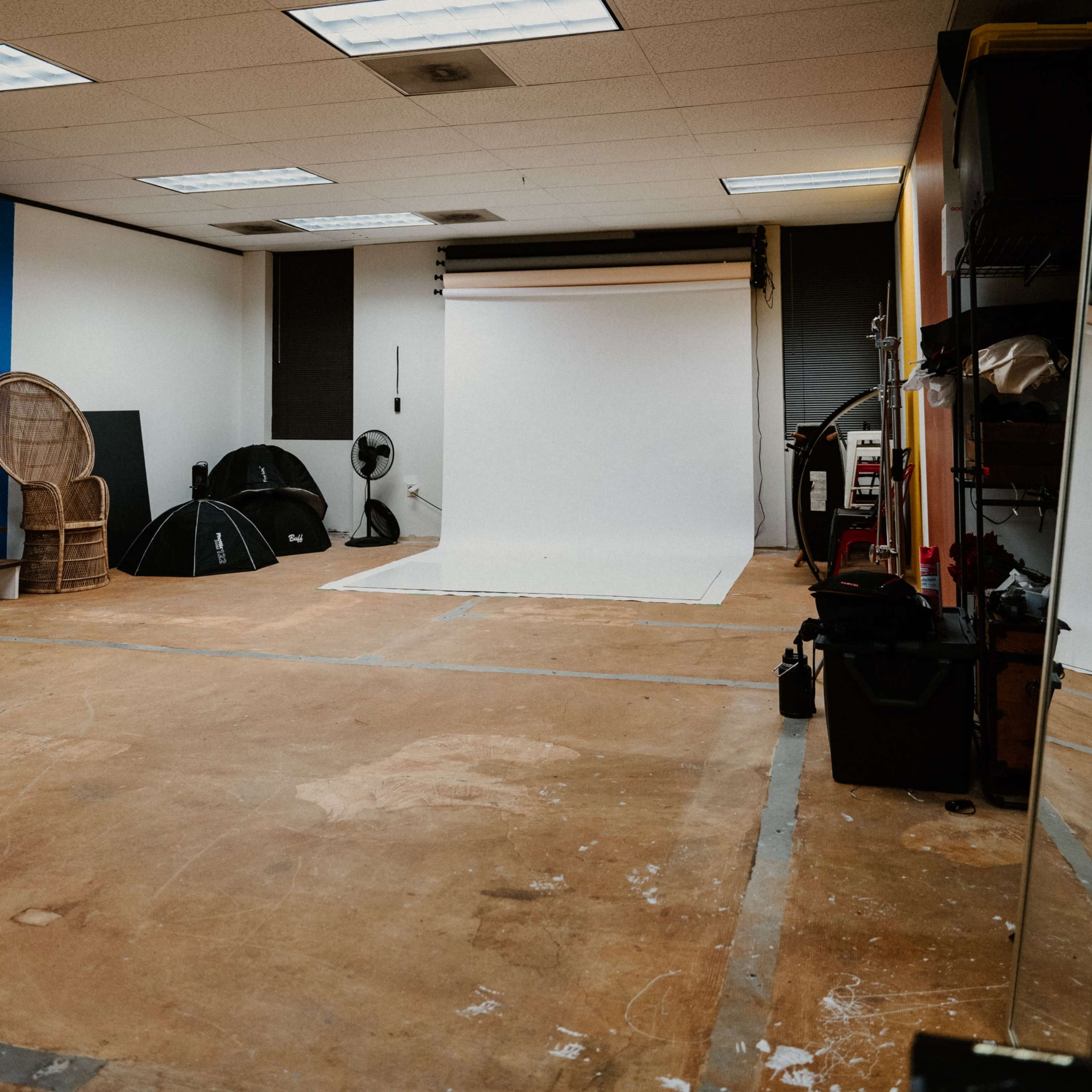 An empty photography studio with a white backdrop, scattered equipment, and chairs arranged along the walls.
