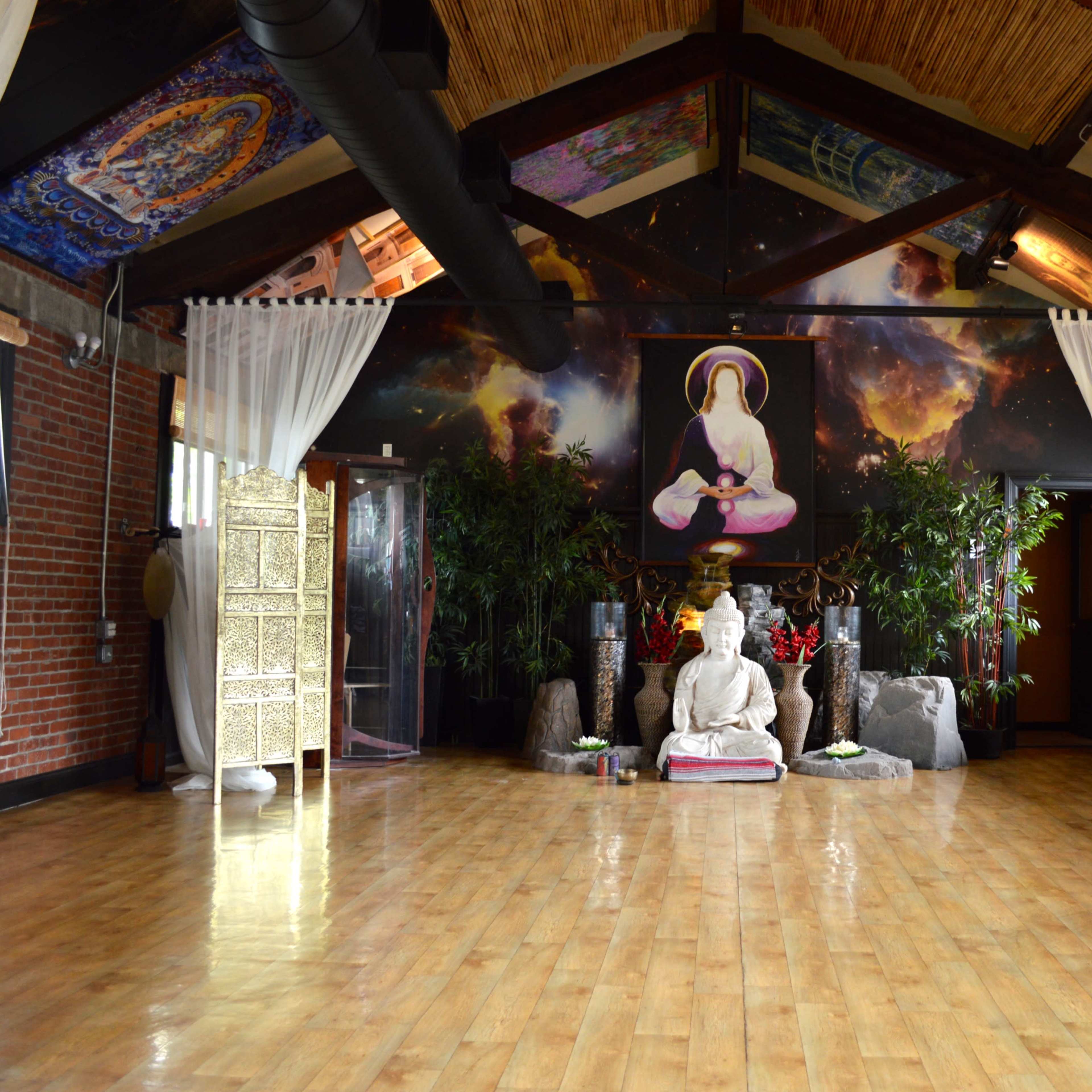 The scene depicts a spacious, well-lit yoga studio featuring a central stone statue of a meditating figure surrounded by plants, decorative elements, and murals on the walls.