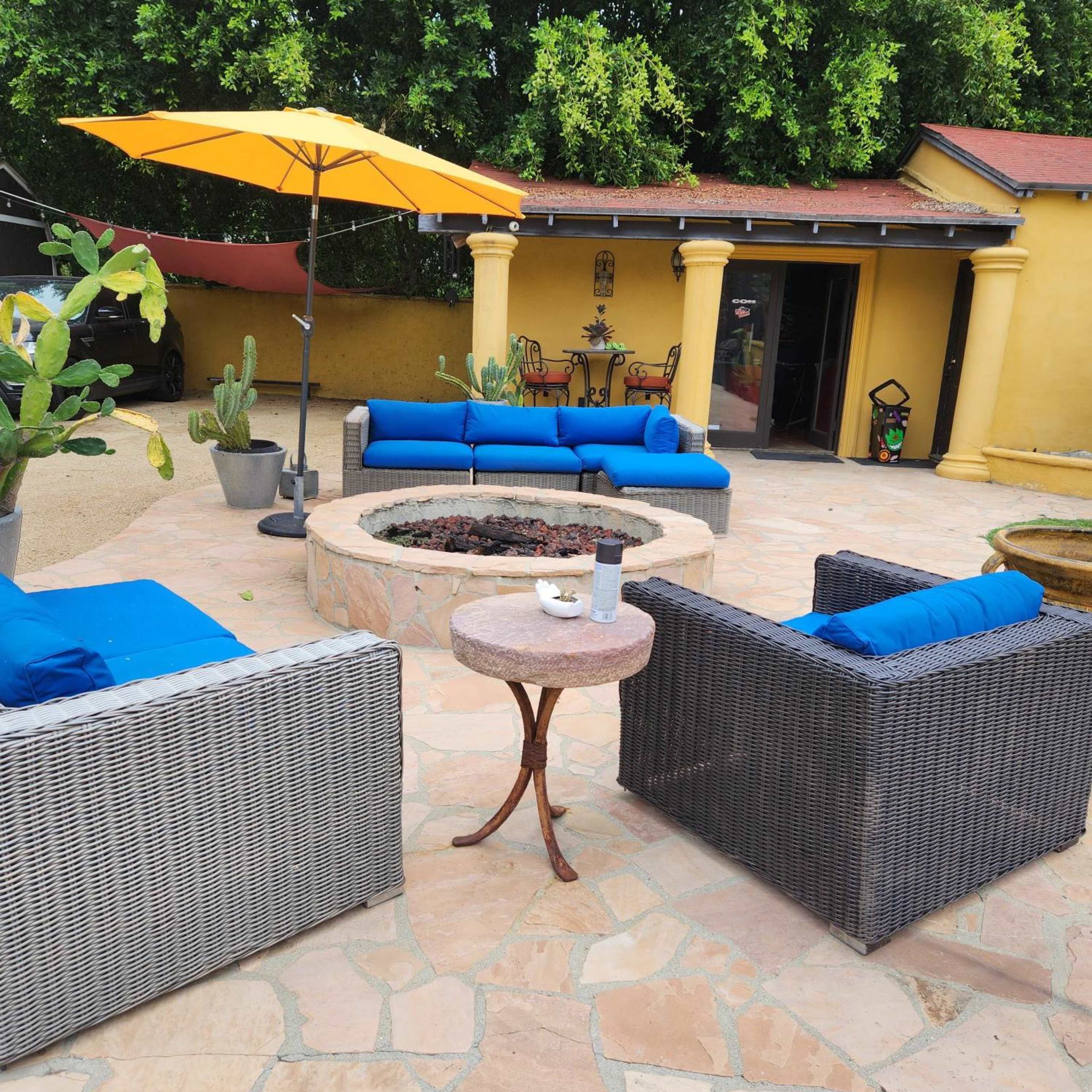 A patio features a circular fire pit surrounded by four outdoor seating arrangements, accented by an umbrella and potted plants.