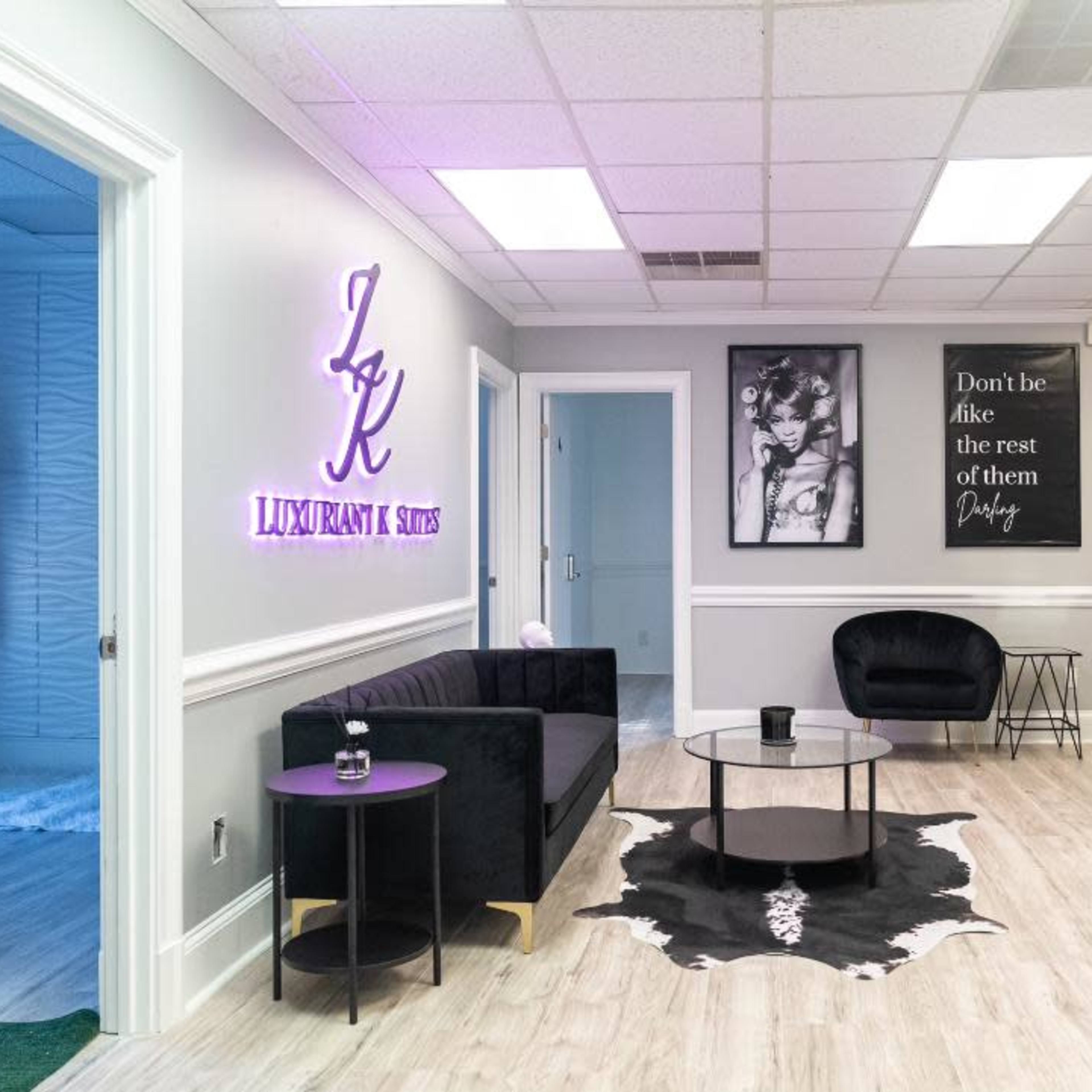 A modern waiting area with a black sofa, a round coffee table, and framed artworks on the walls, featuring a neon sign displaying "LUXURIANT & STYLES."