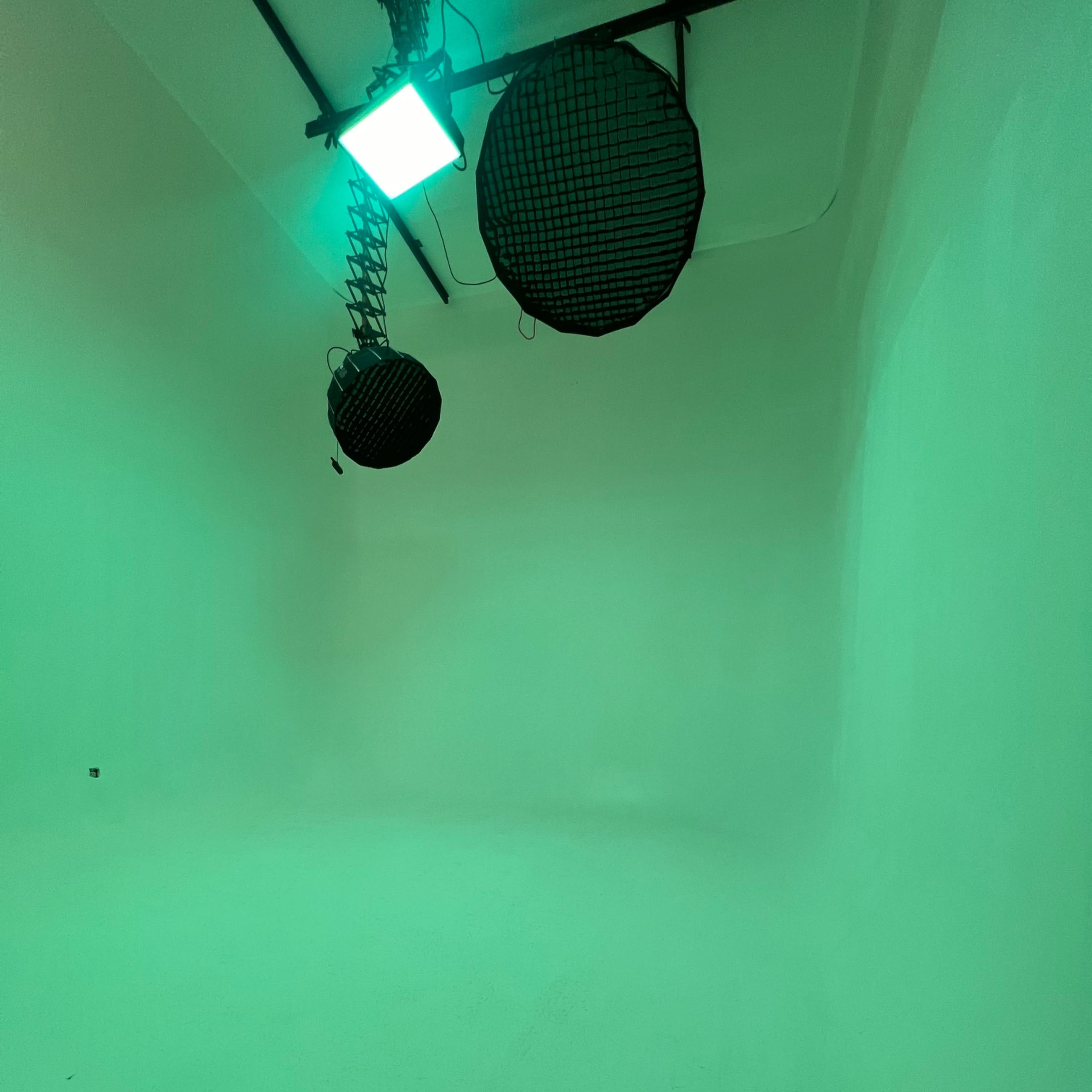 A minimalist studio space with two black lights hanging from the ceiling and green lighting illuminating the white walls.