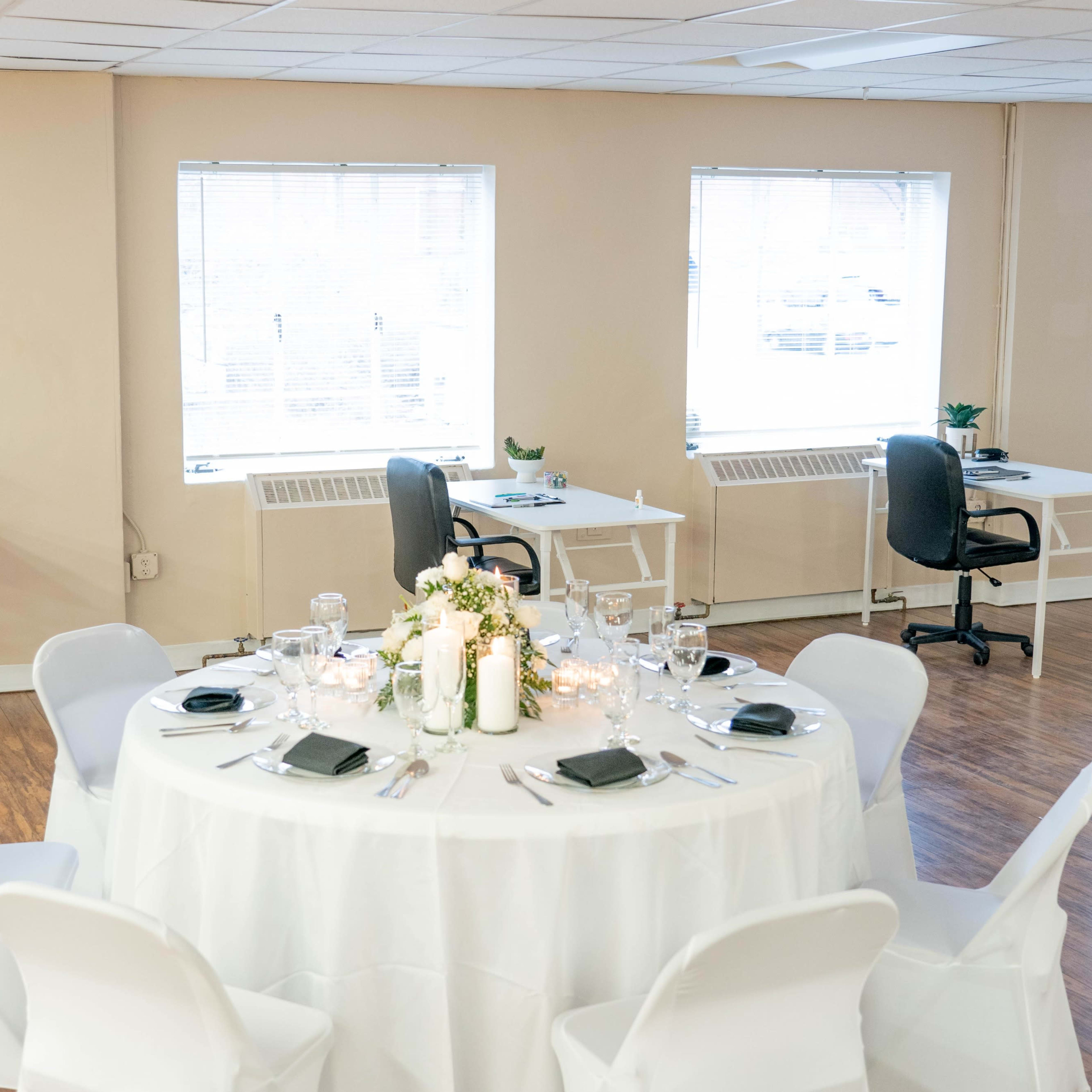 A round dining table is set with dishes and a floral centerpiece, surrounded by white chairs in a well-lit room with additional desks and chairs.