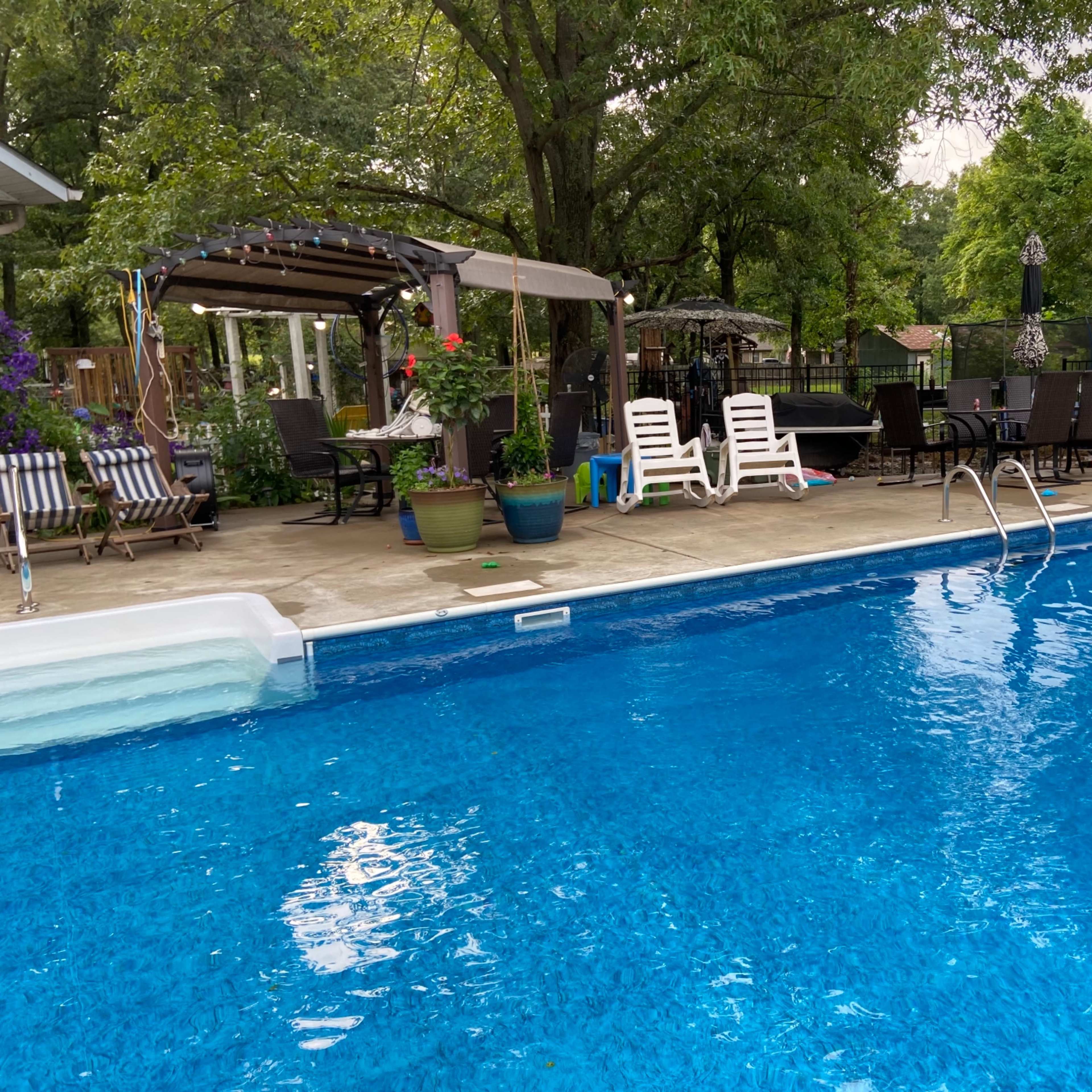 A clear blue swimming pool surrounded by lounge chairs, potted plants, and a shaded seating area with a pergola.