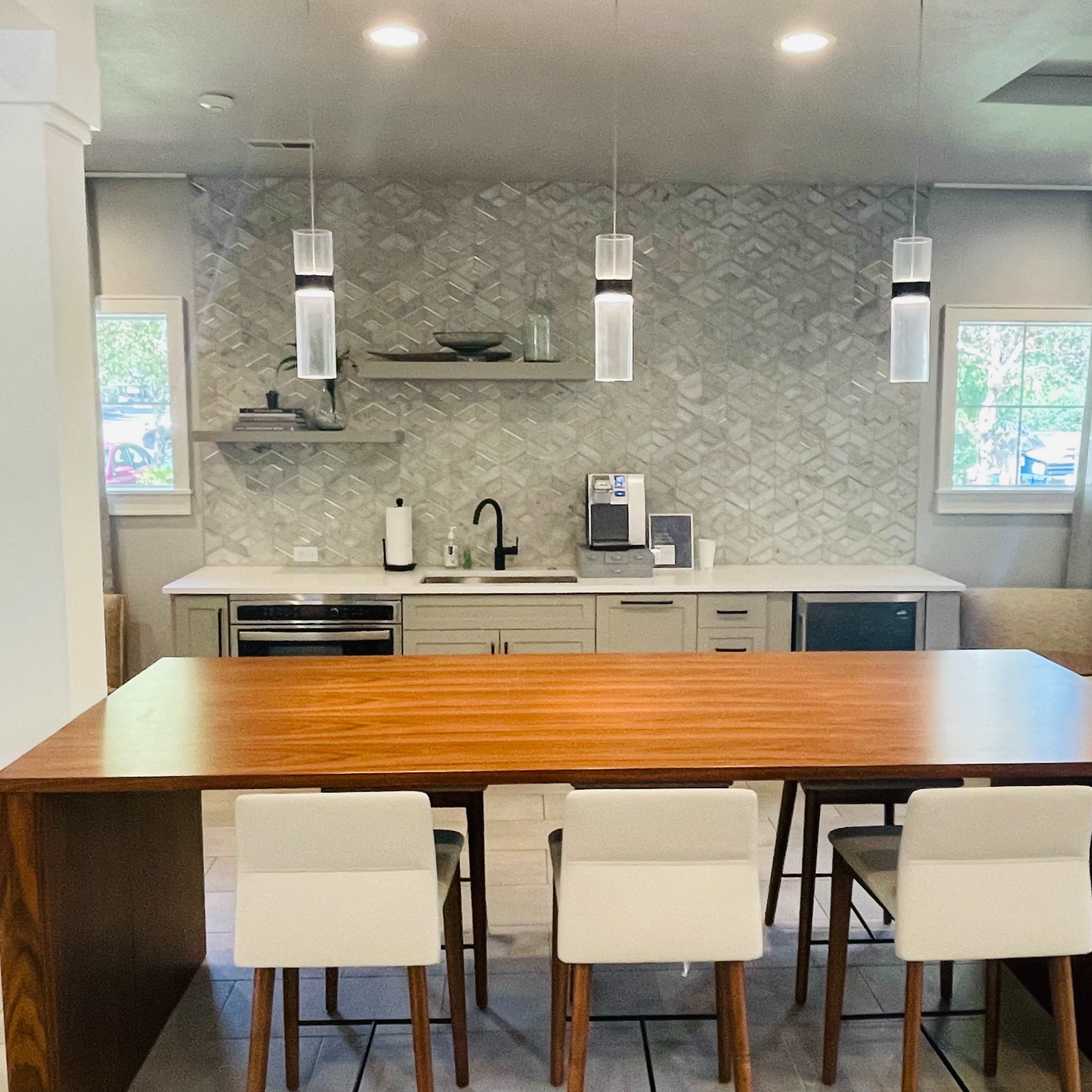 A modern kitchen area with a wooden table and white bar stools, featuring a herringbone-patterned backsplash and appliances against a light-colored wall.