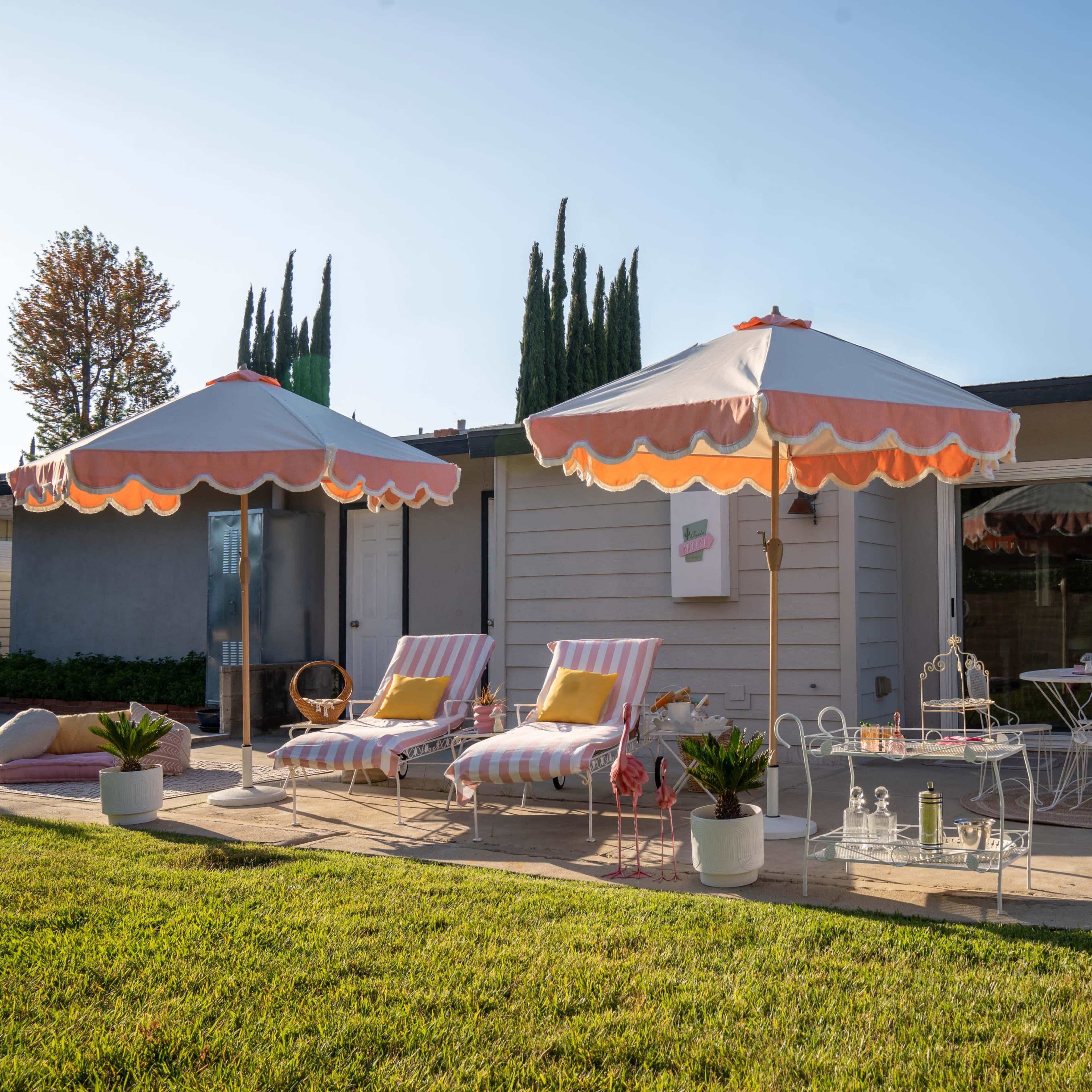 A backyard with two striped lounge chairs and matching umbrellas beside a small glass table, surrounded by potted plants and a manicured lawn.