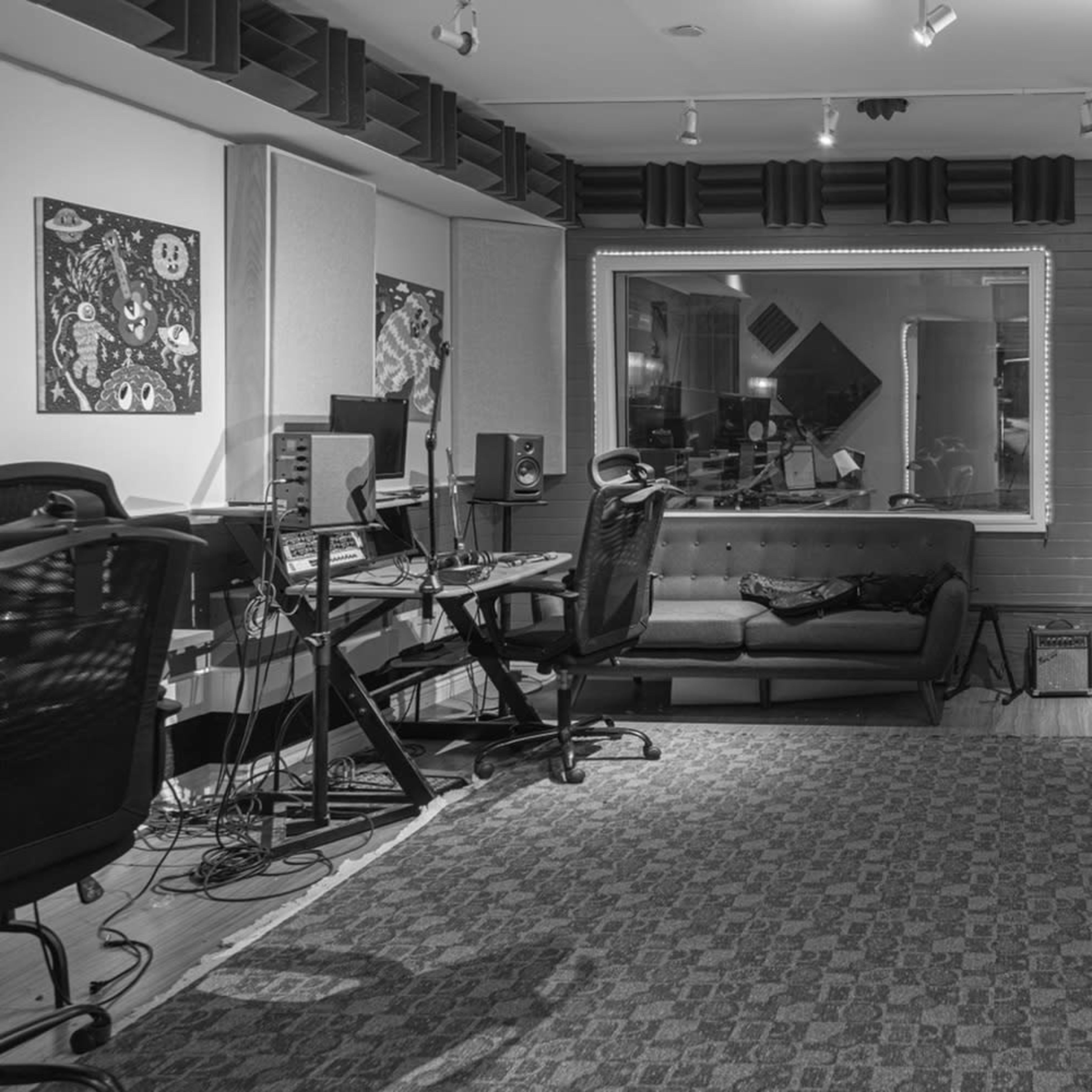 A music recording studio with desks, computer equipment, and a large window overlooking an isolated seating area.