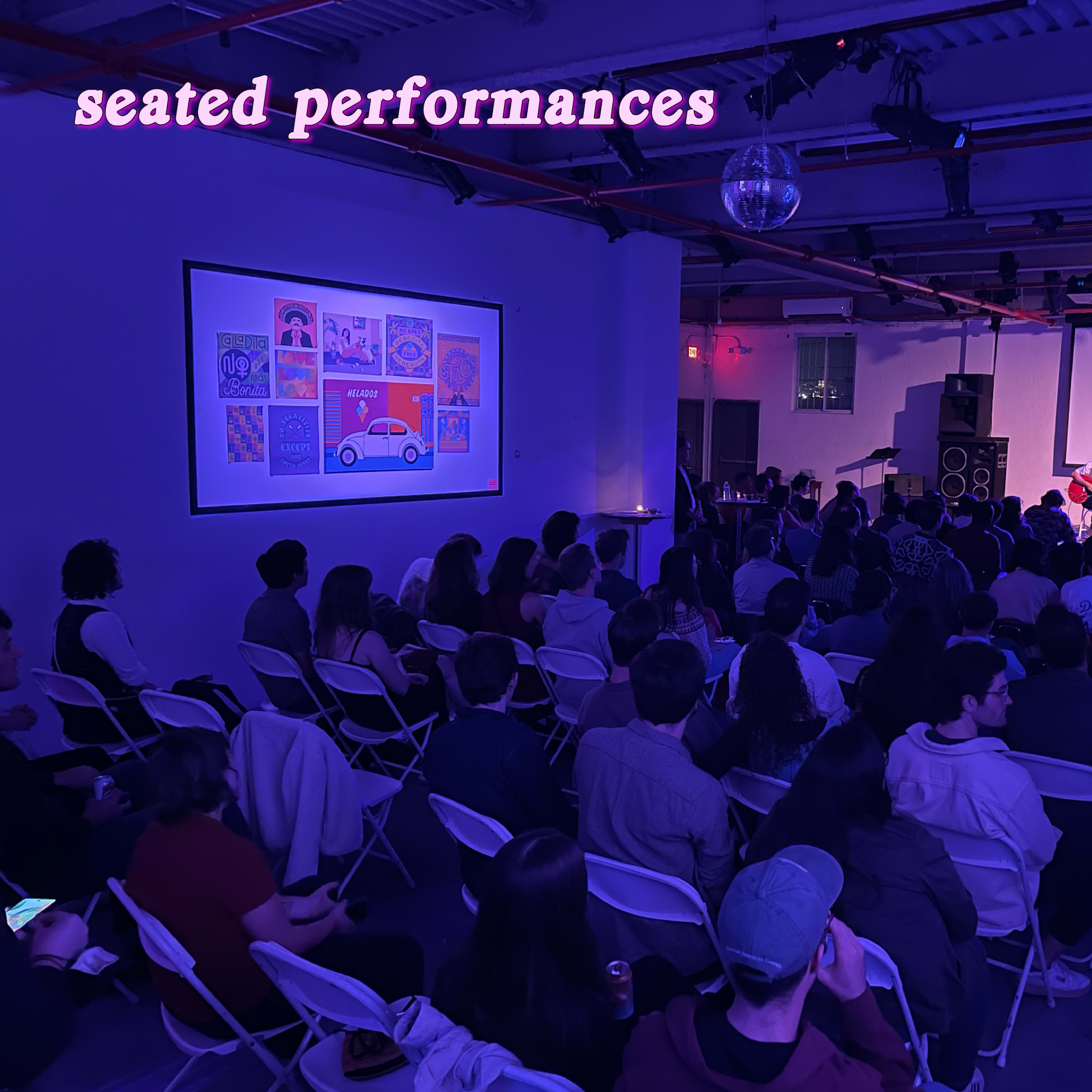 A crowd is seated in an event space, listening to a discussion while a colorful artwork projection is displayed on the wall.