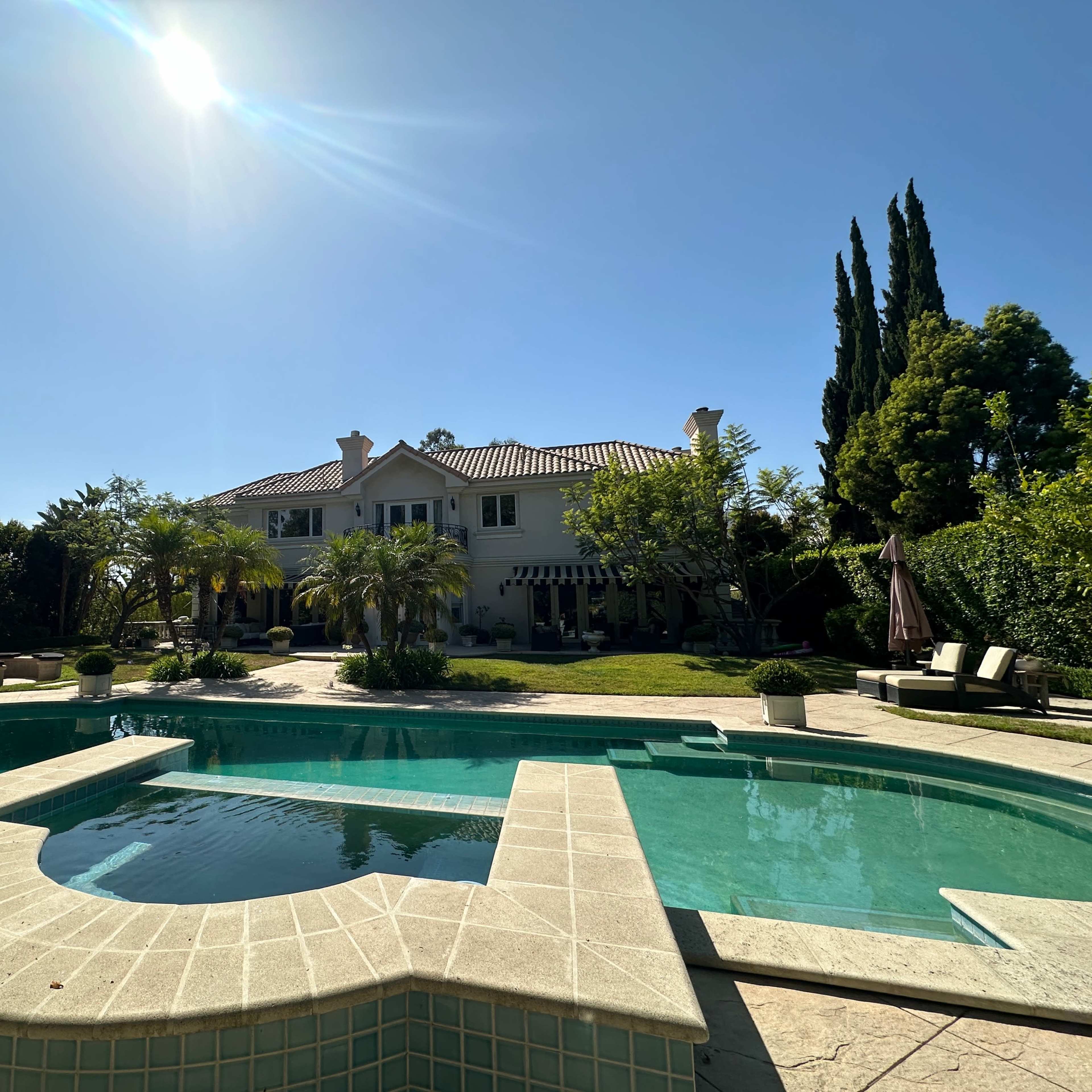 A spacious backyard features a swimming pool with a spa, surrounded by palm trees and a large two-story house in the background.