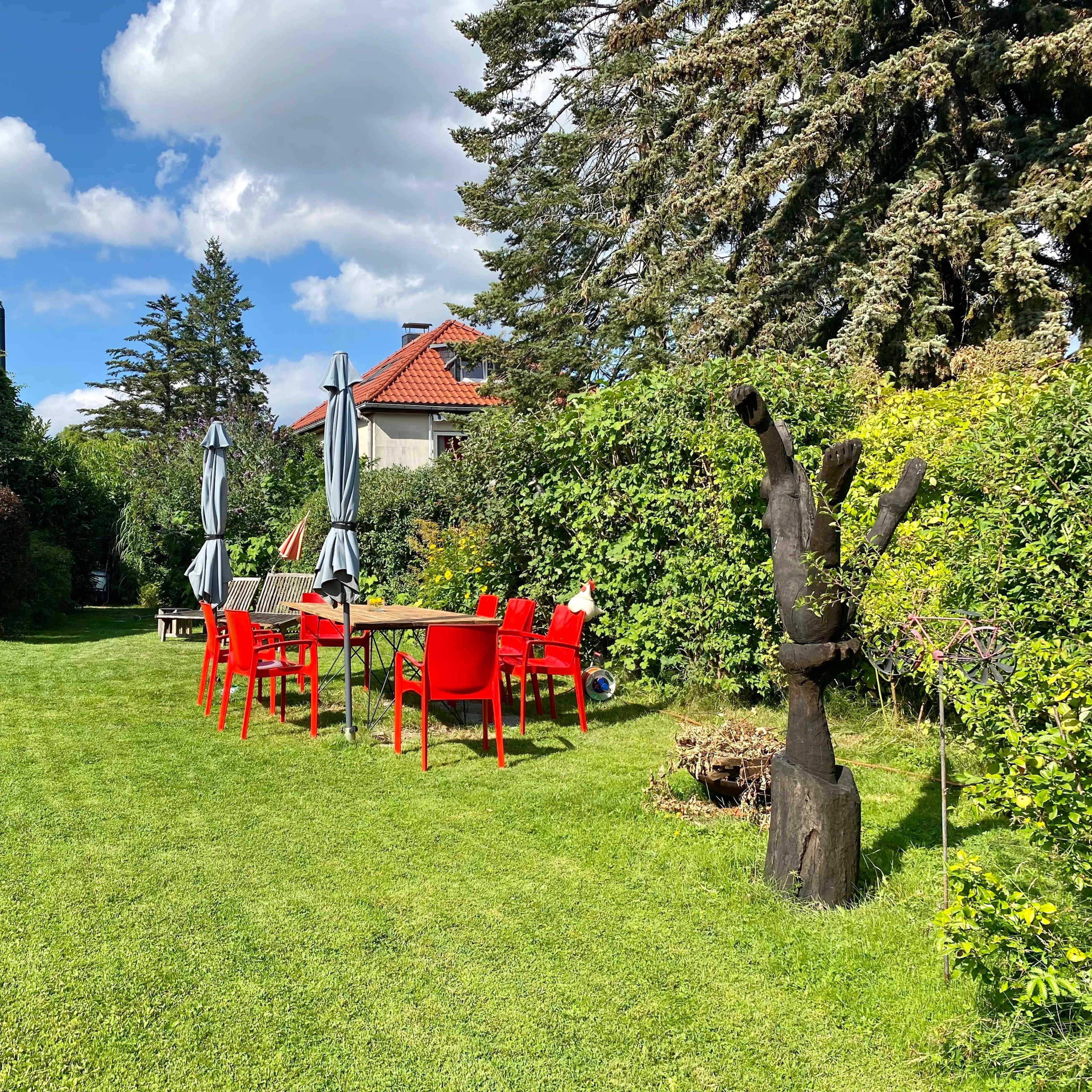 A bright green lawn features a red outdoor dining set surrounded by trees and shrubs under a partly cloudy sky.