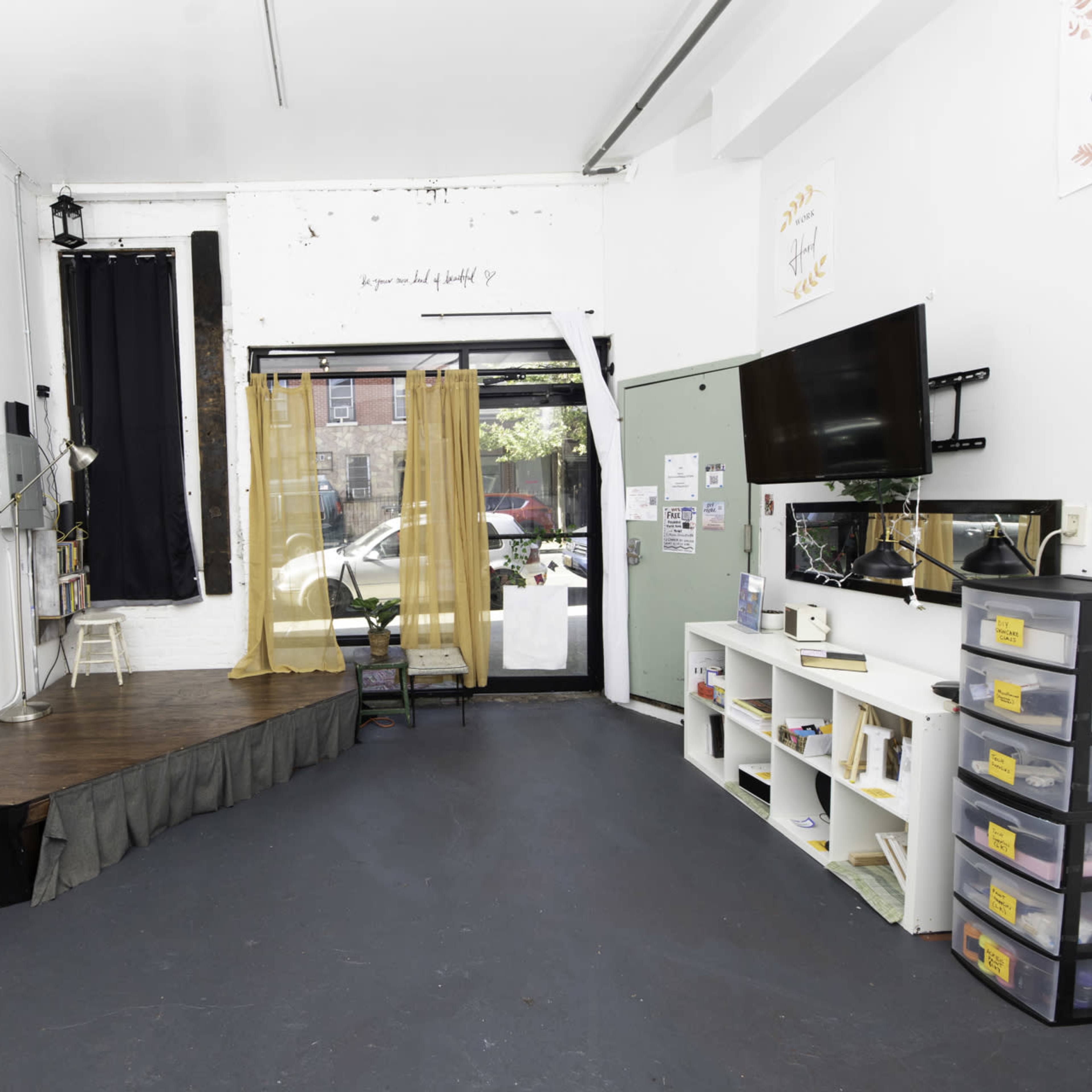 A small, empty room with a wooden stage, a black curtain, a television mounted on the wall, and several storage bins.