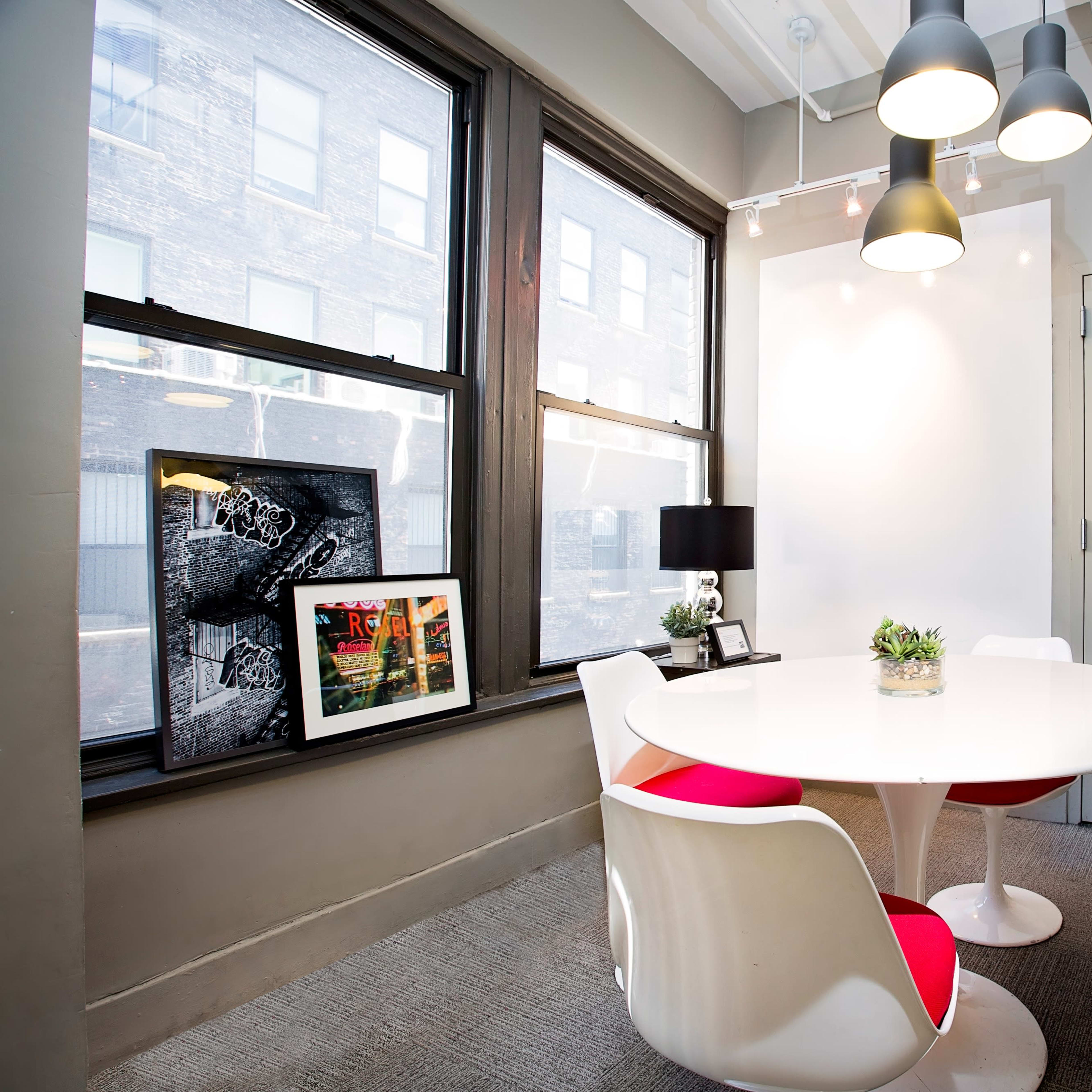 A modern dining area features a round white table surrounded by four white and red chairs, with large windows and a plant centerpiece.