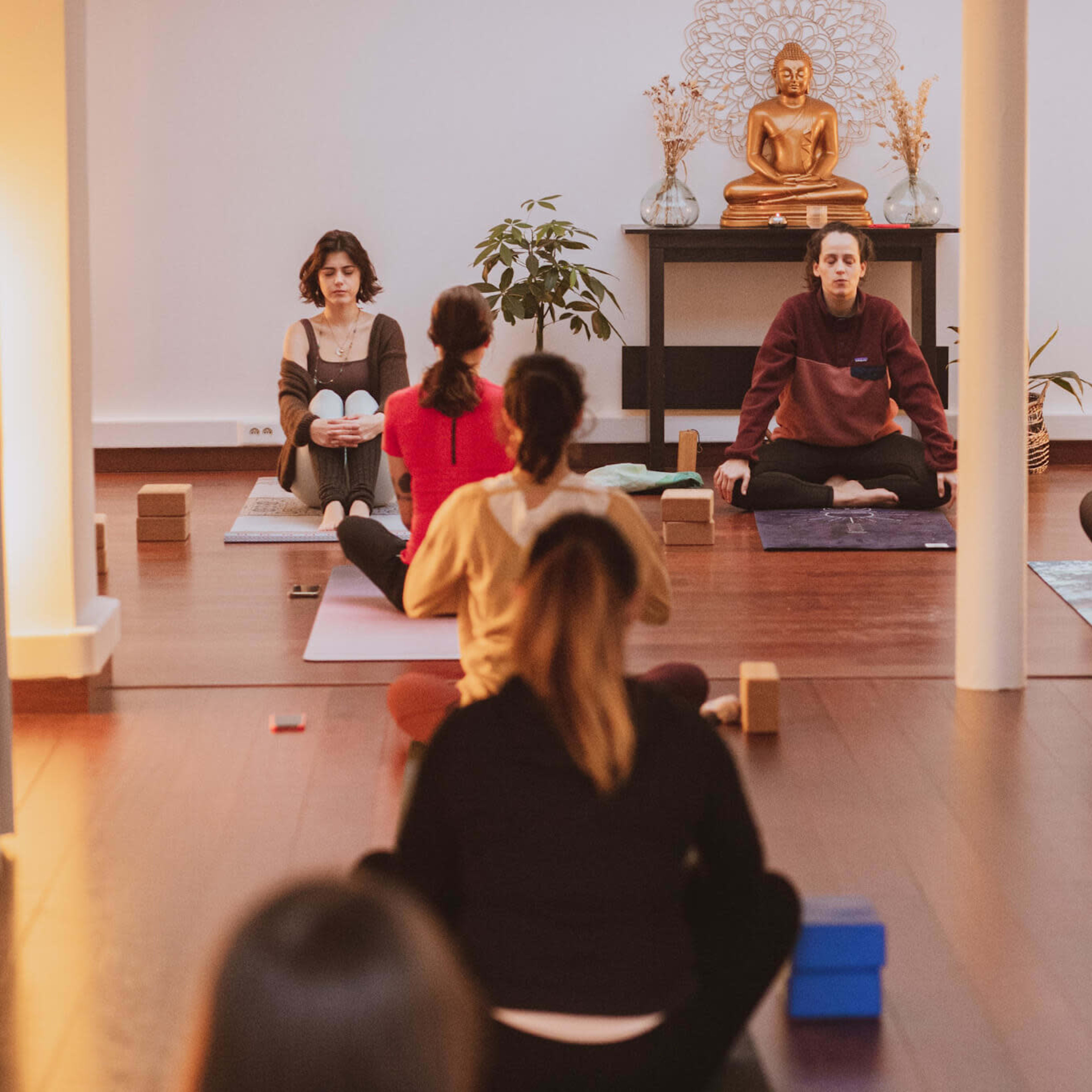 A group of people sits on yoga mats in a peaceful studio, facing a statue of a Buddha surrounded by soft lighting and plants.