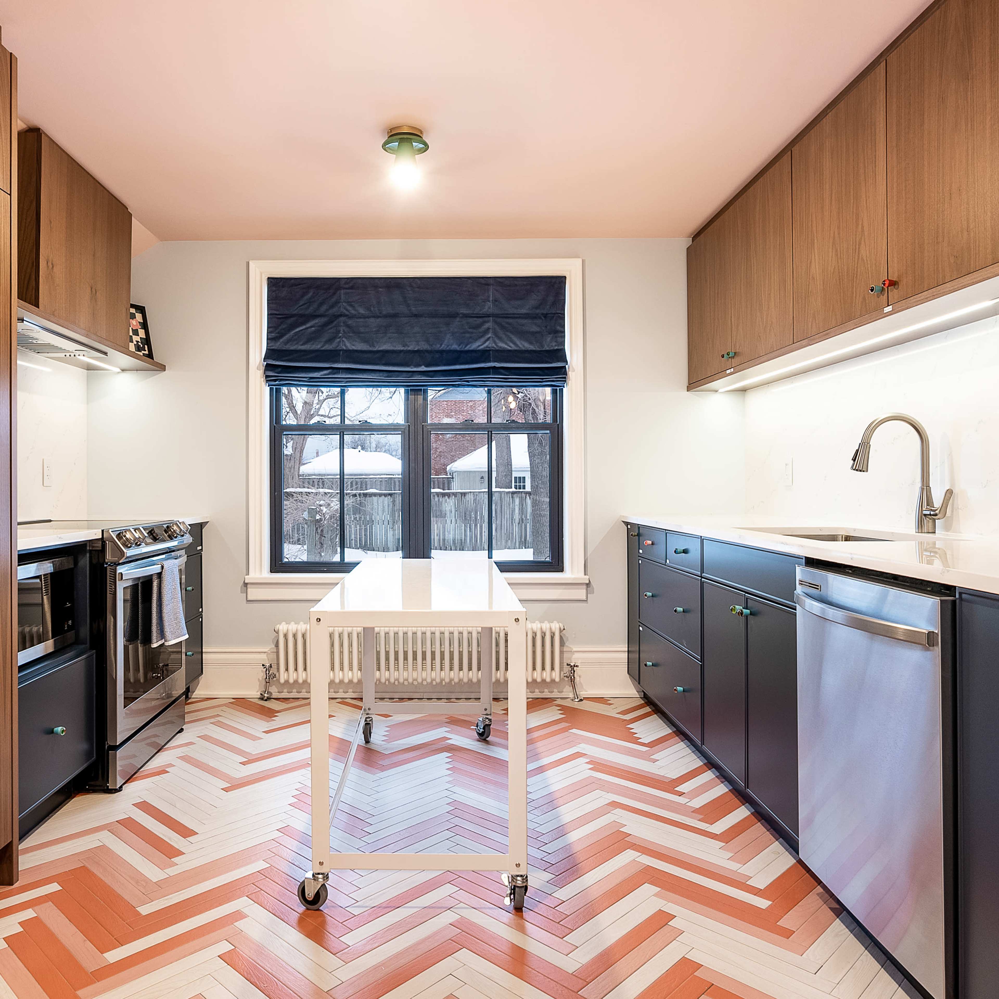 A modern kitchen features stainless steel appliances, dark cabinetry, and a chevron-patterned floor with a central table.