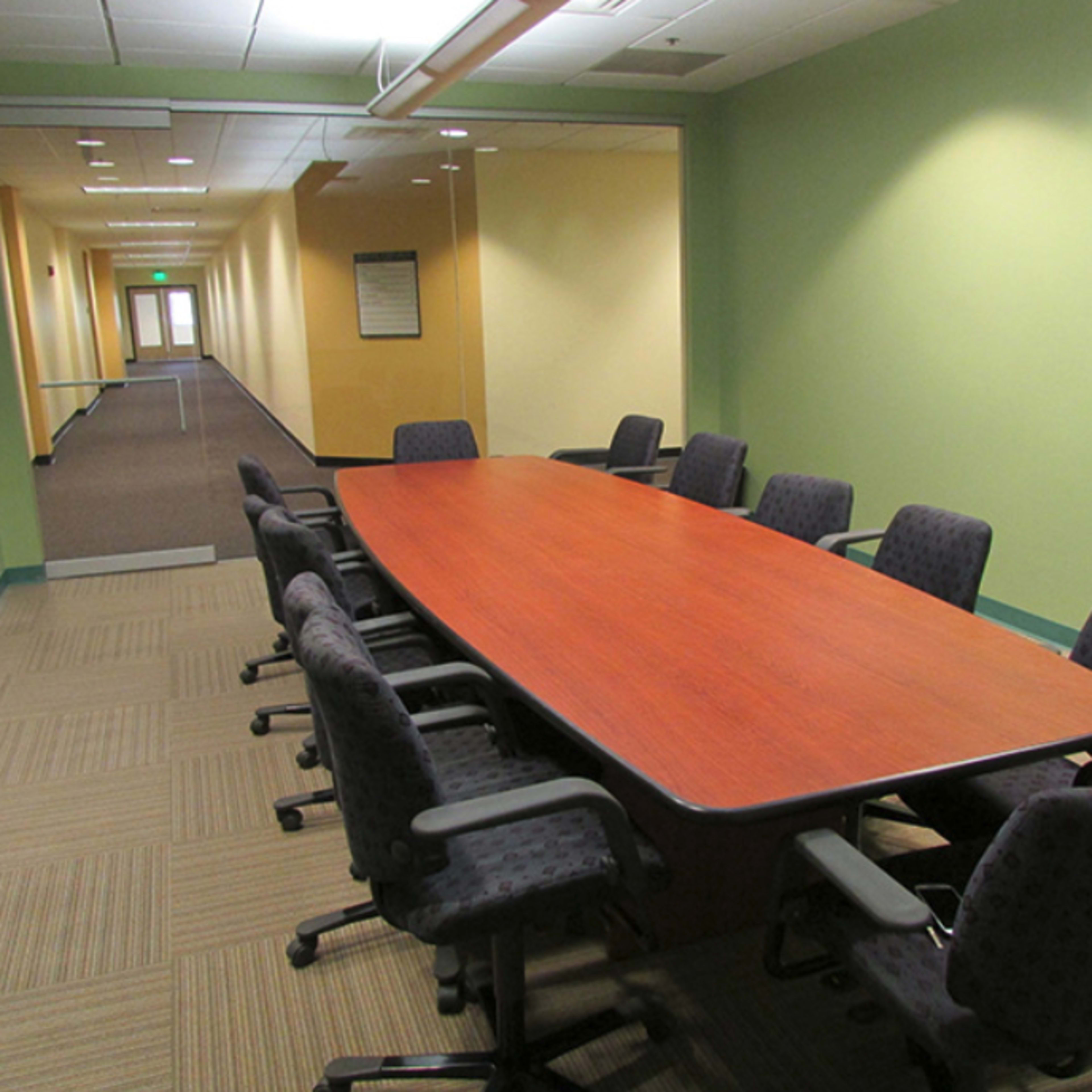 A long, wooden conference table is surrounded by eight upholstered chairs in a brightly lit office space.