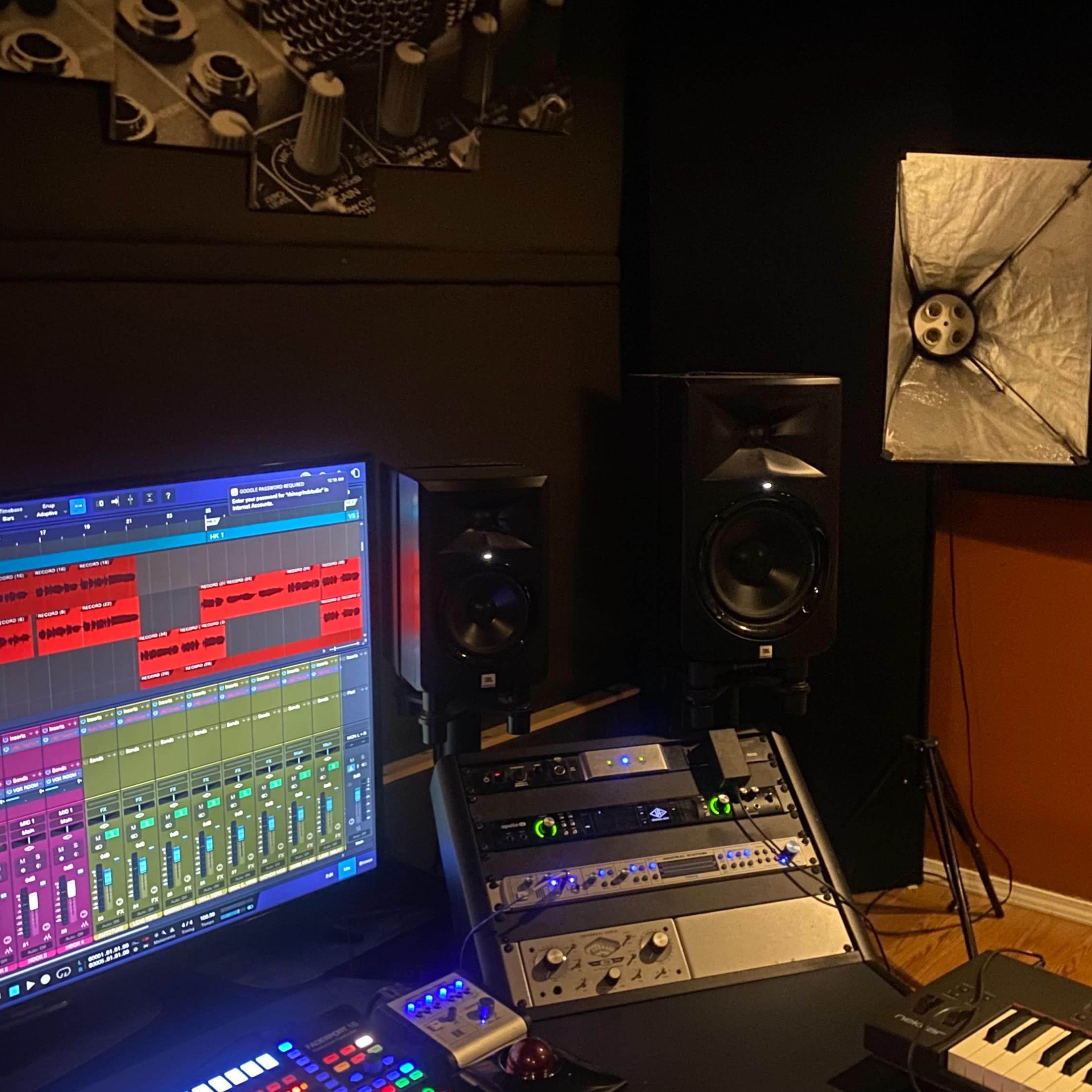A recording studio setup features a large computer monitor displaying audio tracks, mixing equipment, studio speakers, and a keyboard.
