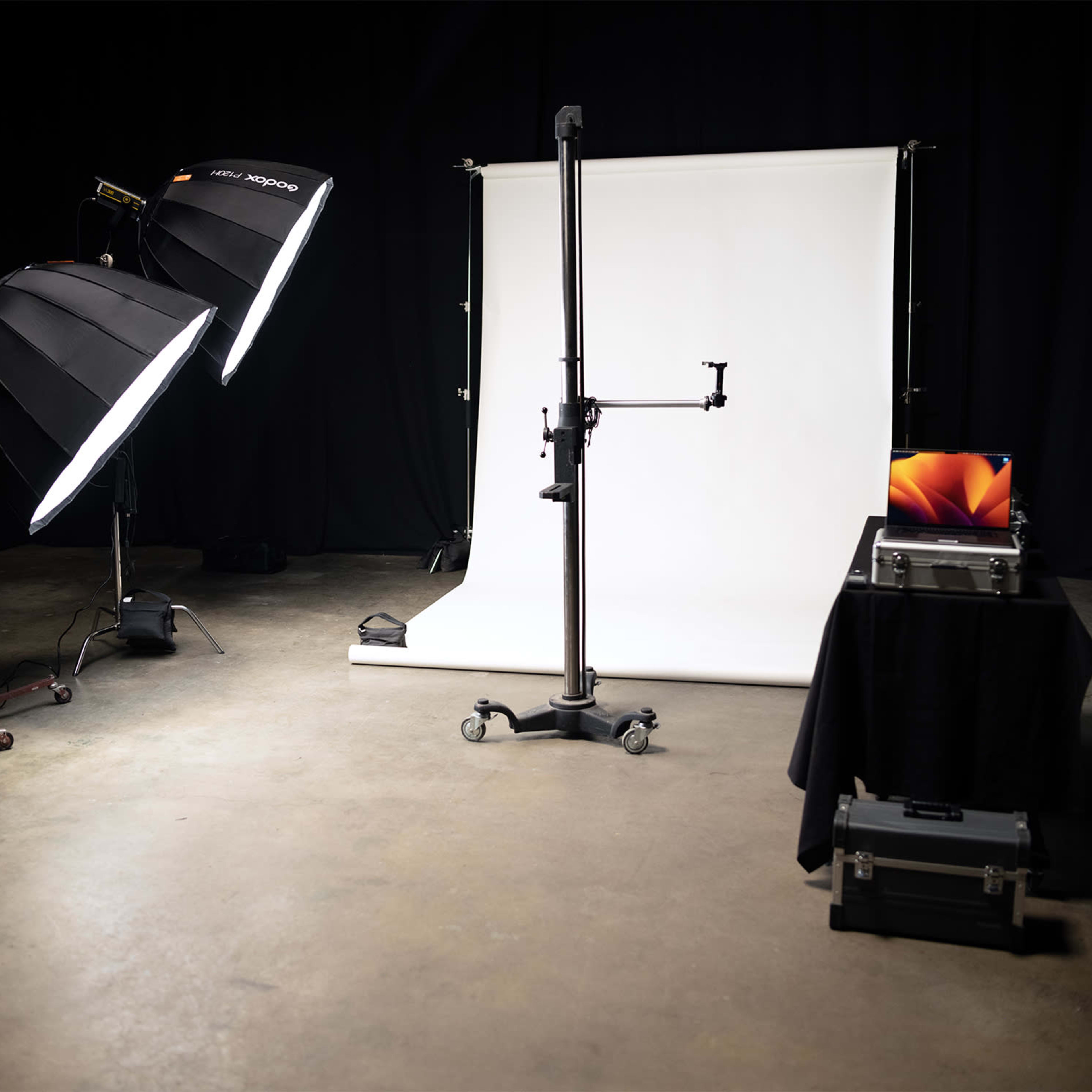 A studio setup with two large softbox lights, a backdrop stand holding a white backdrop, a laptop on a table, and a black case on the floor.