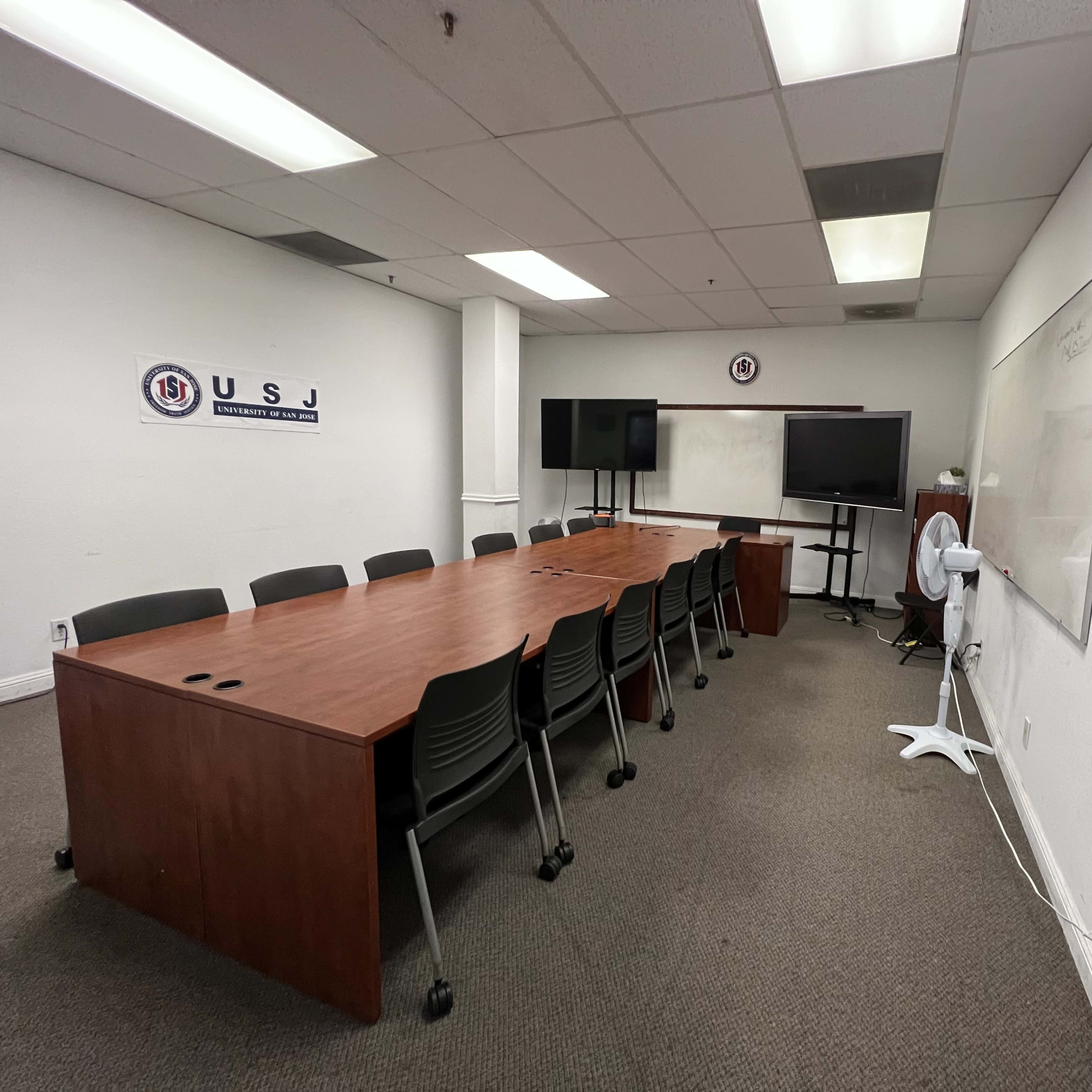 A conference room features a long wooden table surrounded by chairs, with multiple screens and a whiteboard on the walls.