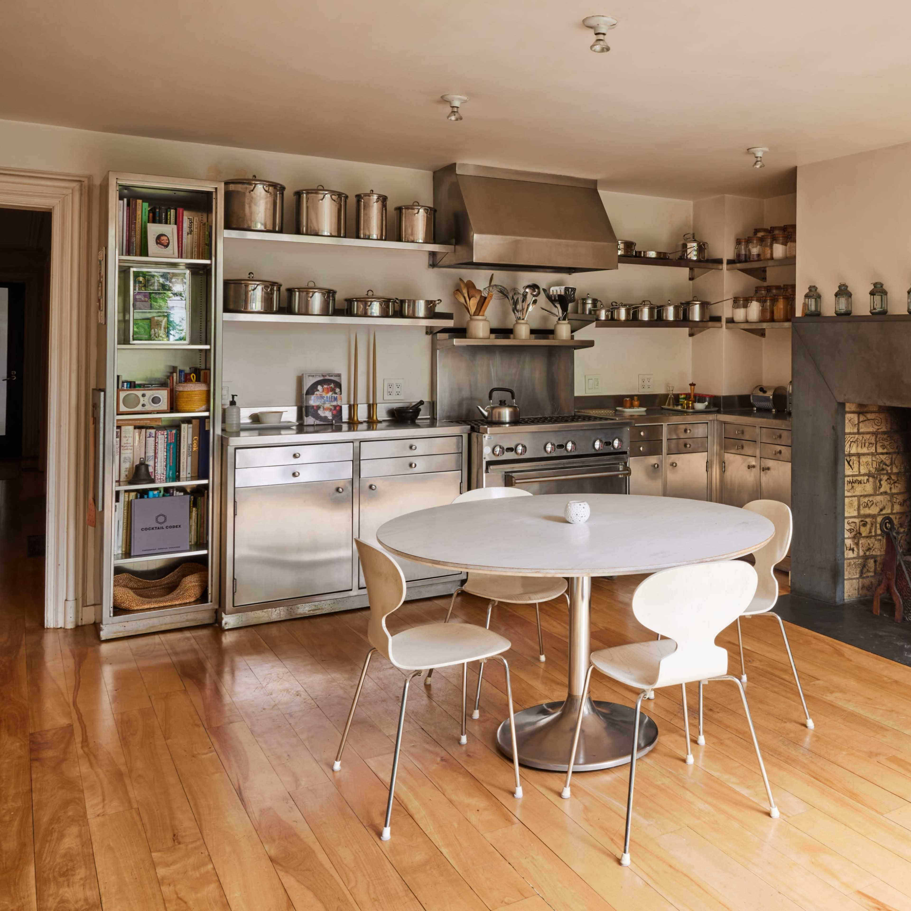 A modern kitchen with stainless steel appliances, shelves filled with jars, and a round table surrounded by four white chairs.