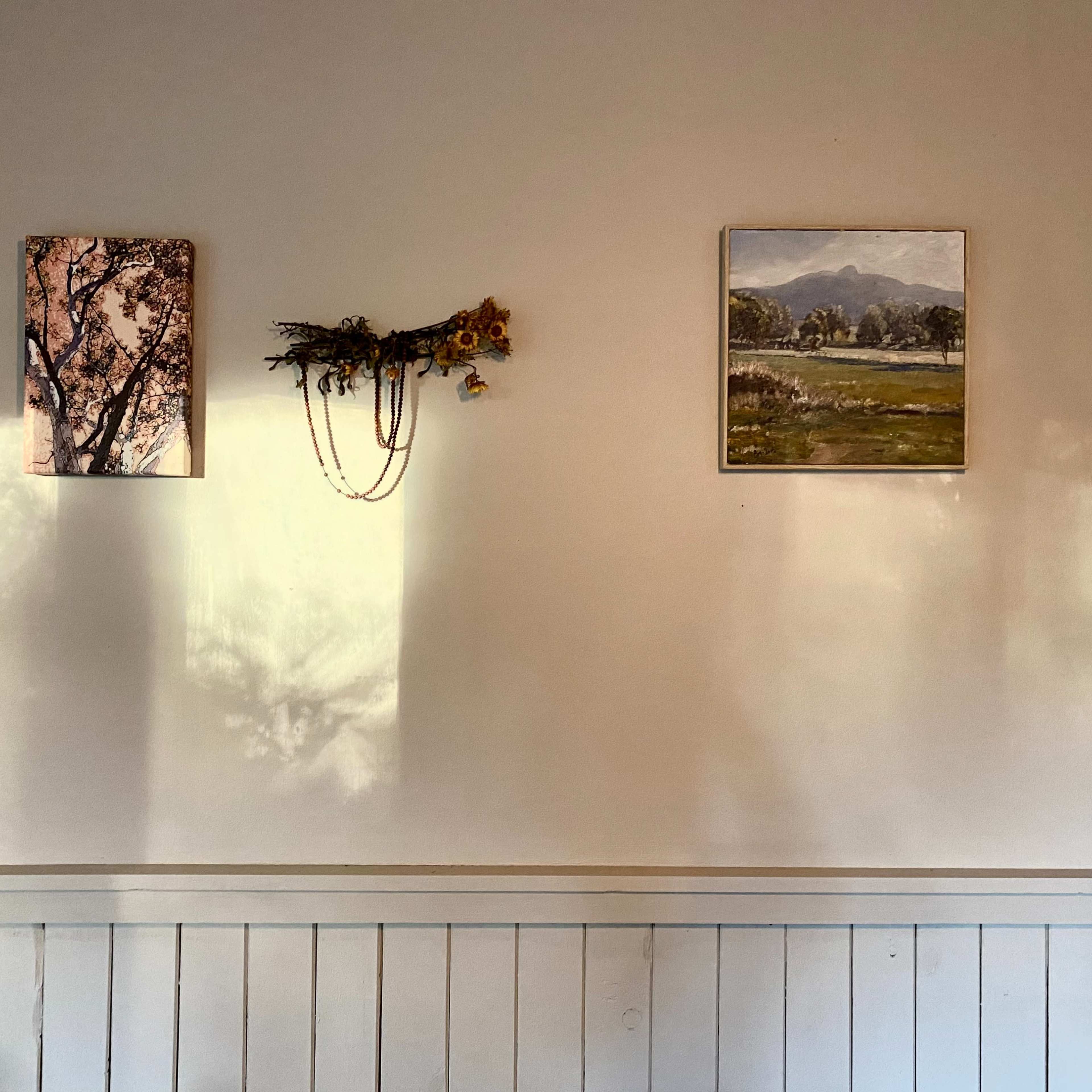 A light-colored wall with two framed pictures and a decorative arrangement of dried flowers, casting shadows.
