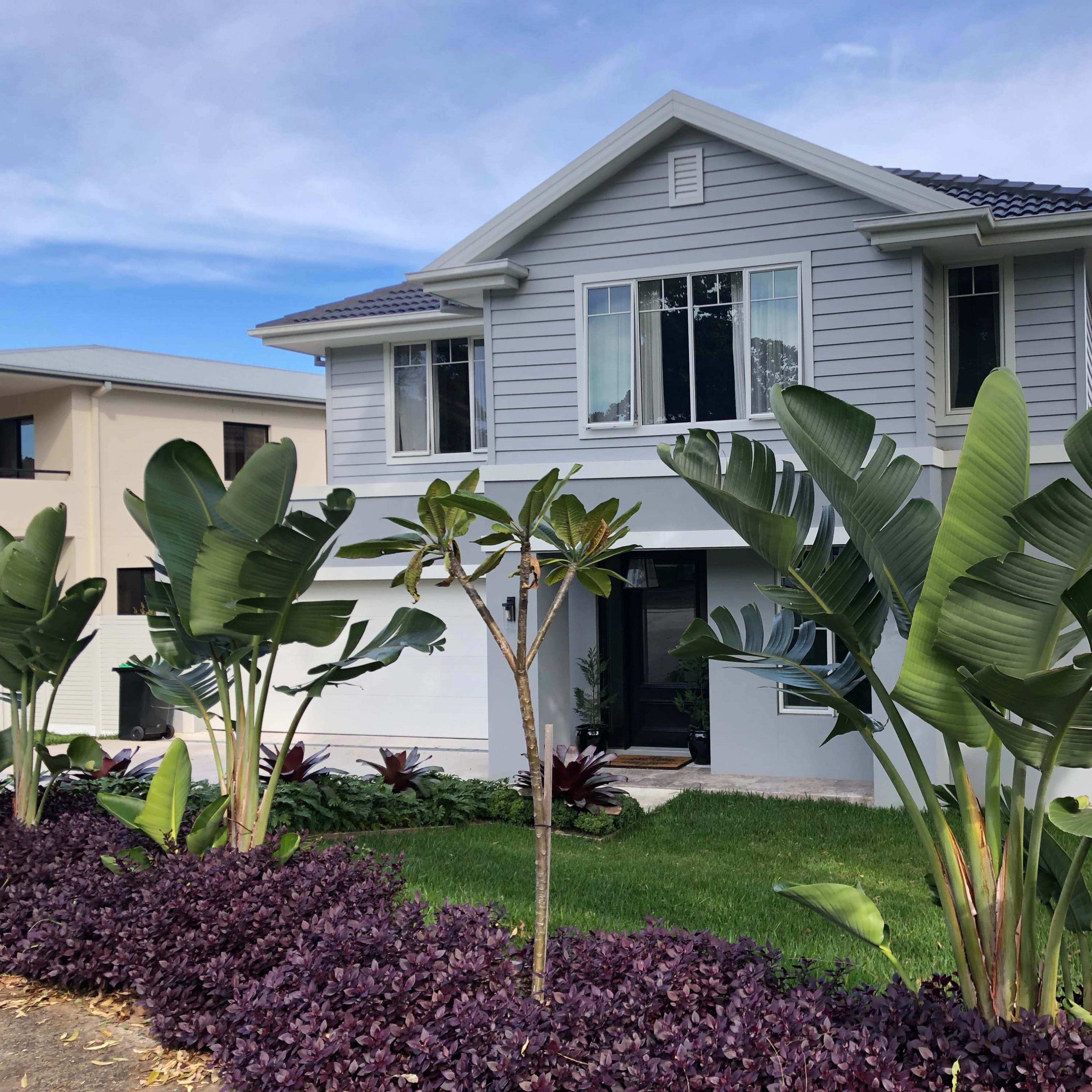 A two-story gray house with large windows is surrounded by tropical plants and a well-maintained garden.