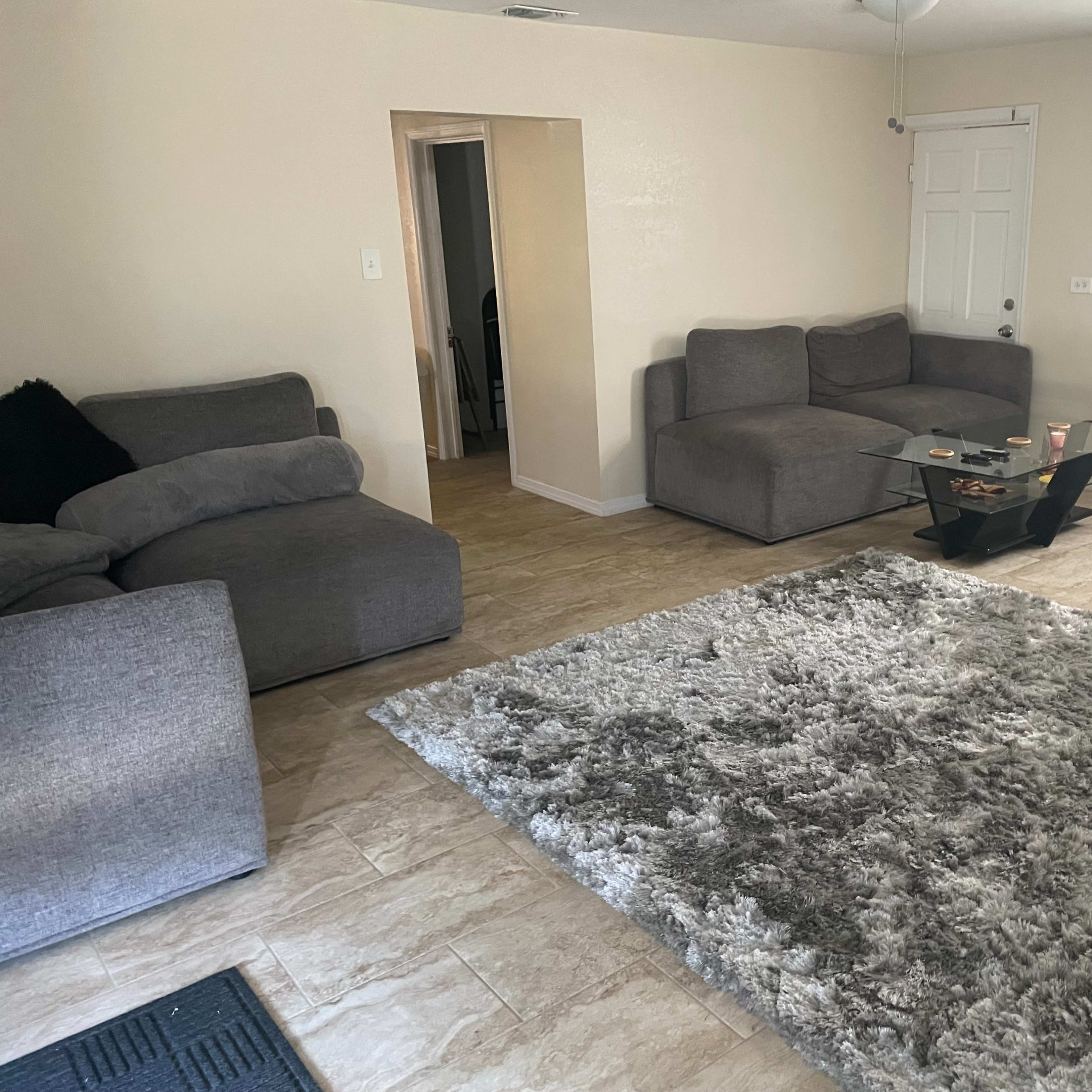 A living room with gray sofas, a glass coffee table, and a large area rug on a tiled floor.
