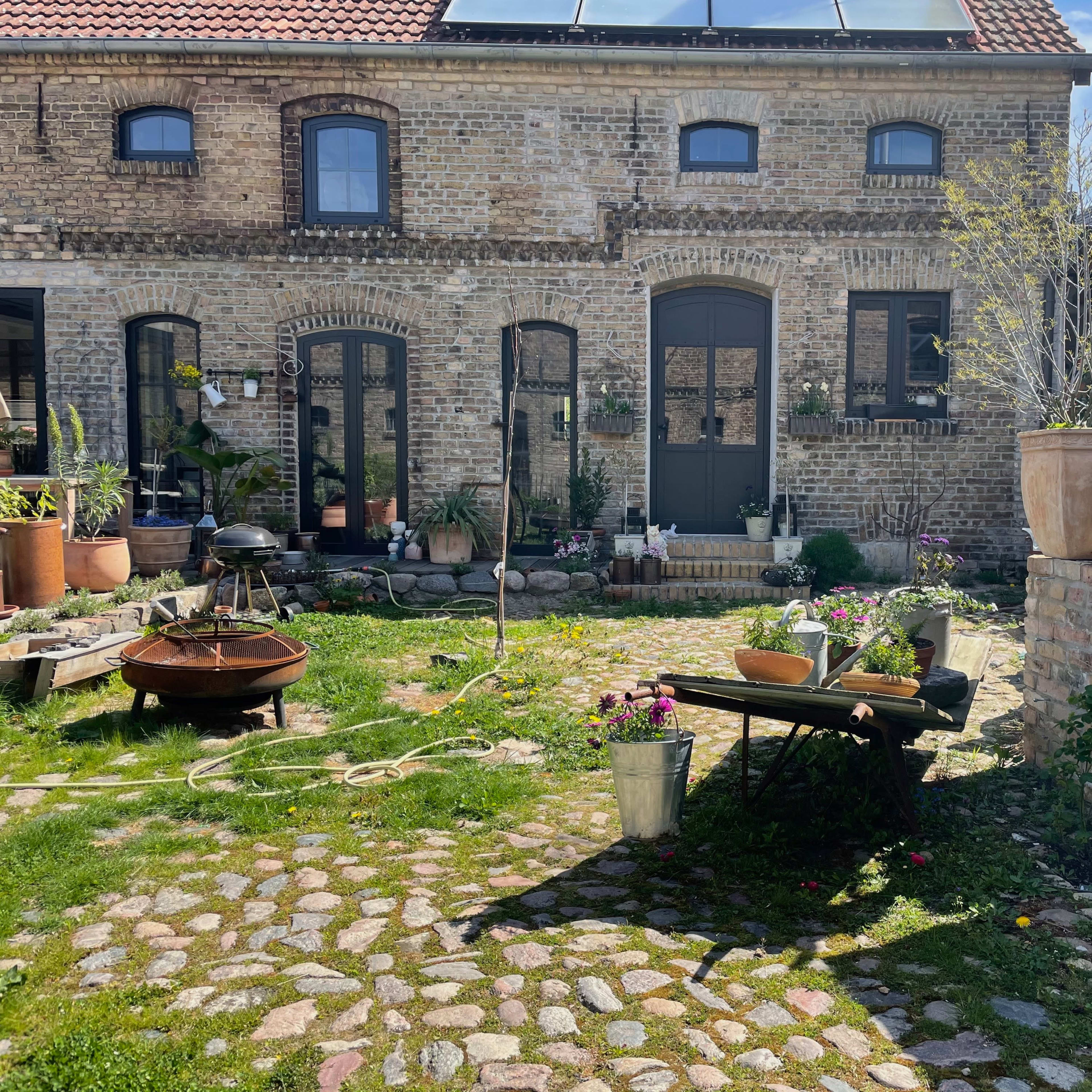 A cobblestone courtyard with a brick building featuring arched windows and a door, surrounded by various potted plants and a table with gardening supplies.