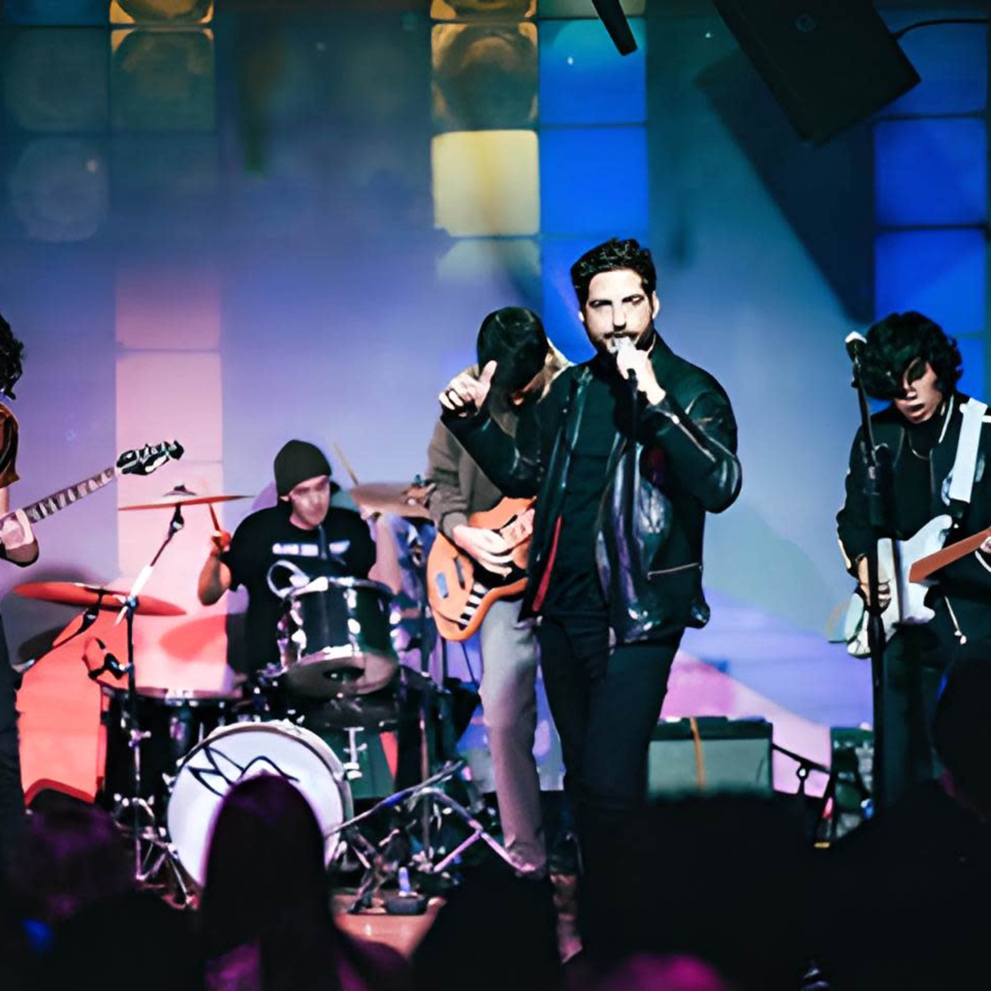 A band performs on stage with a drummer, guitarists, and a lead singer in a colorful lit environment.