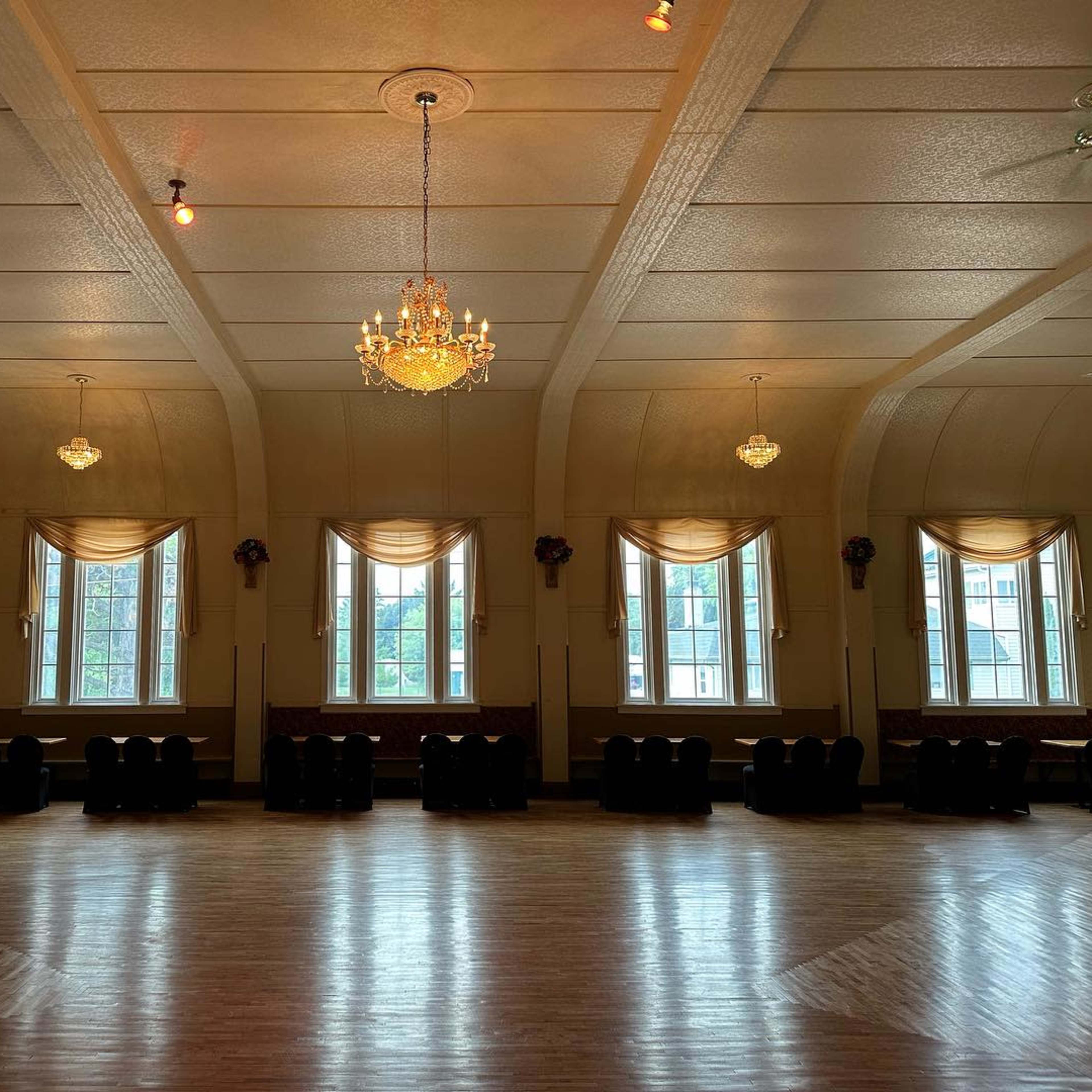A large, empty hall with wooden flooring, adorned with a chandelier and multiple windows dressed with draped curtains.