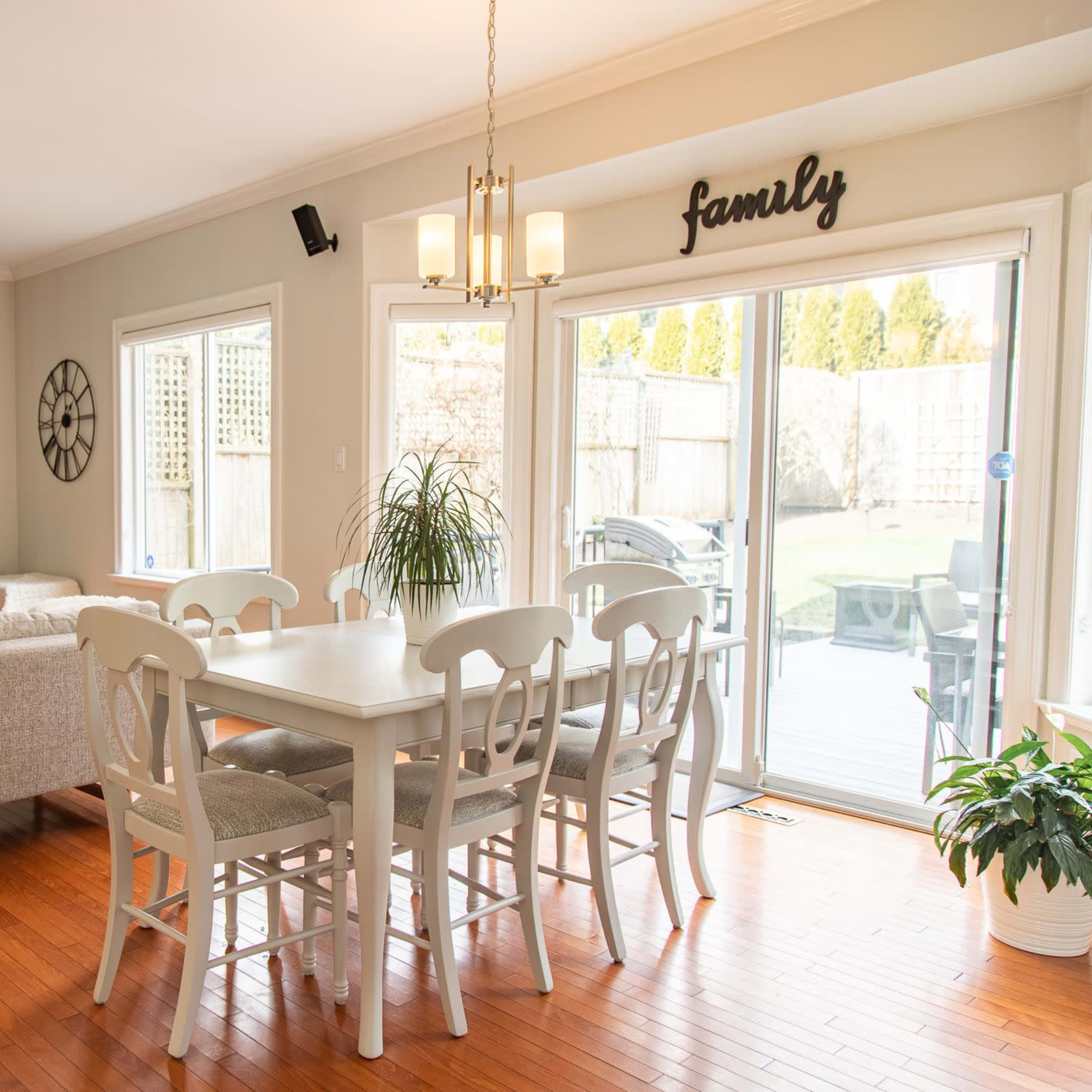 A bright living and dining area with a wooden dining table and chairs, a stone fireplace, and large windows overlooking a backyard.