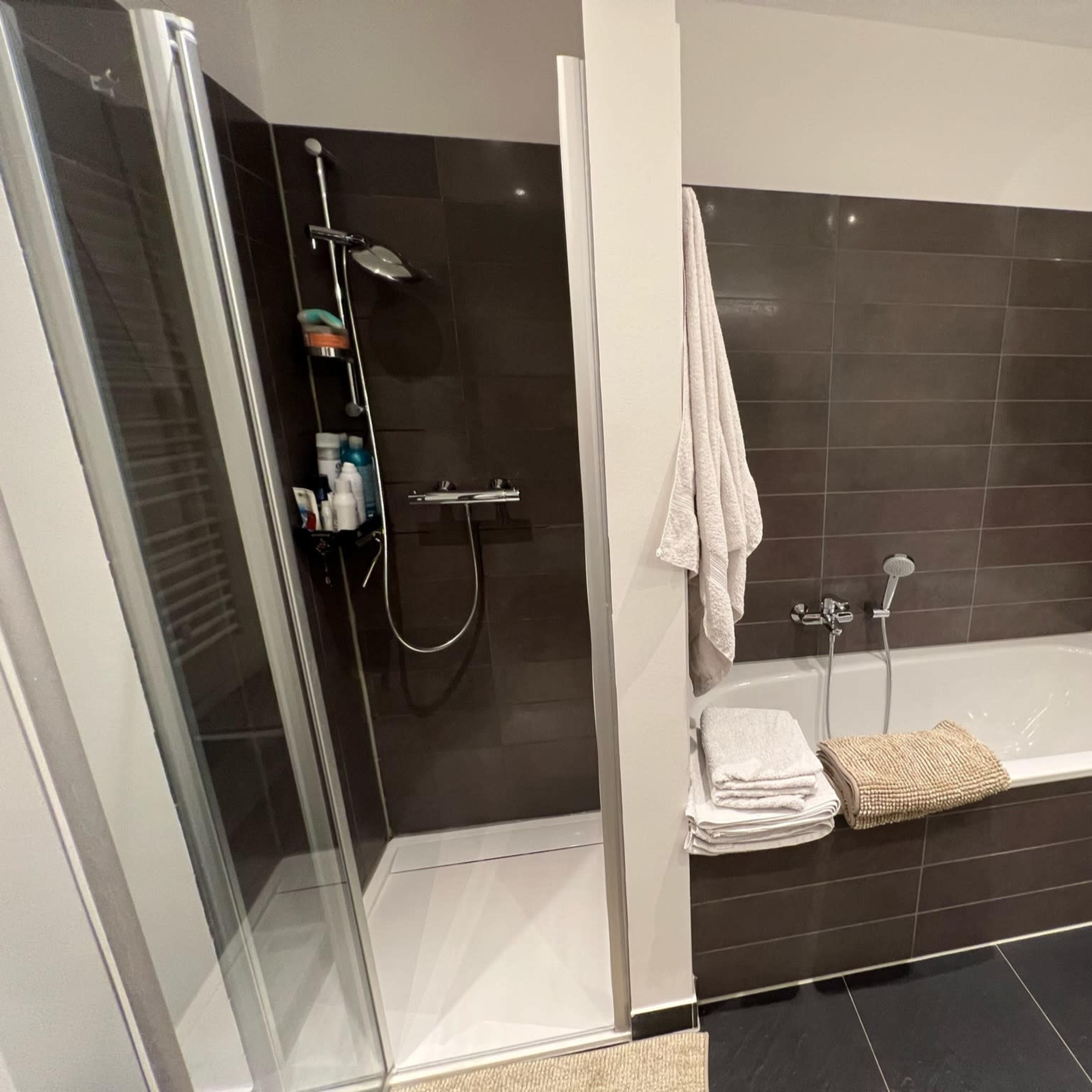 A modern bathroom featuring a shower area on the left and a bathtub with a towel and toiletries on the right.
