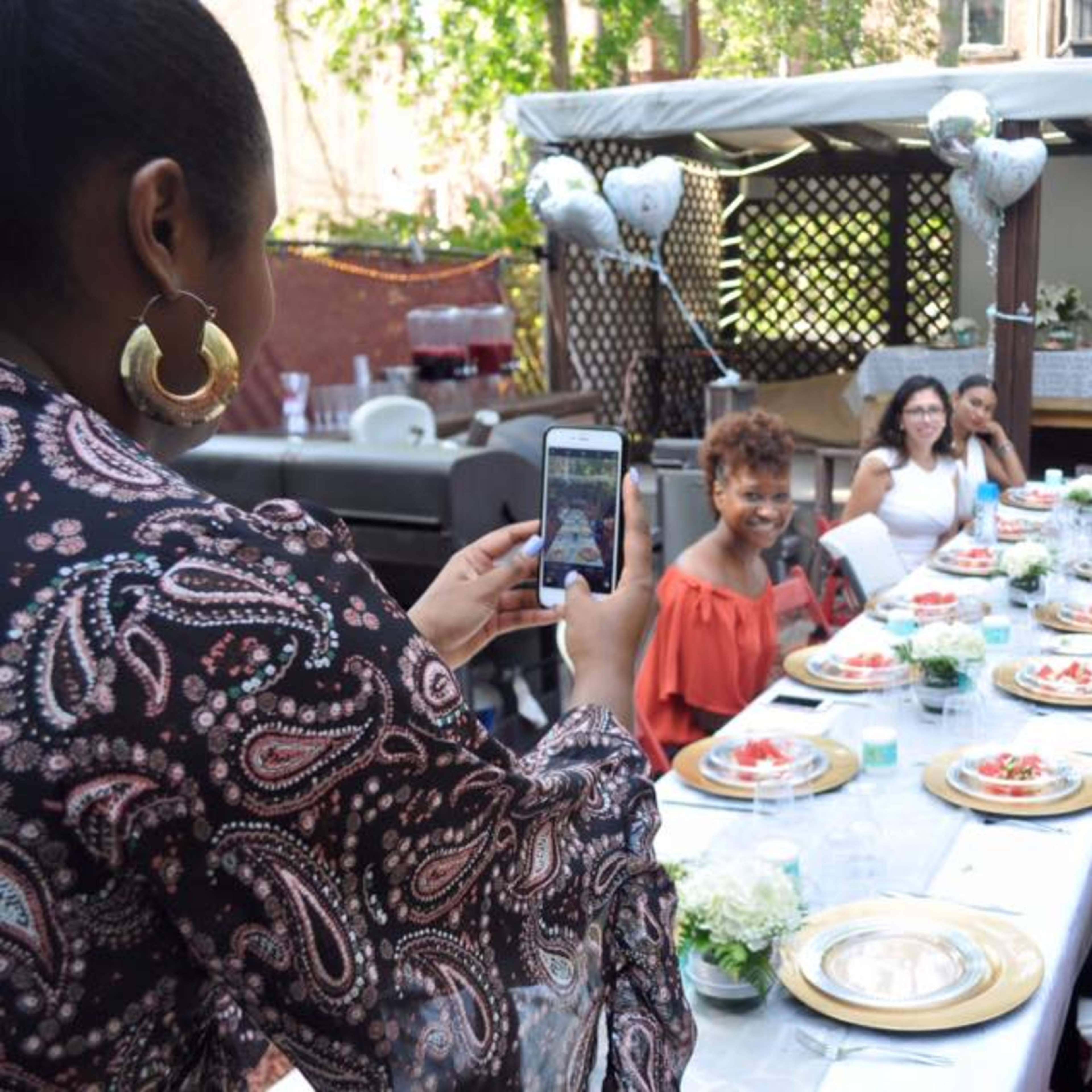 A group of six women are seated around a table set for a meal in an outdoor setting, while one woman stands and takes a photo of them.